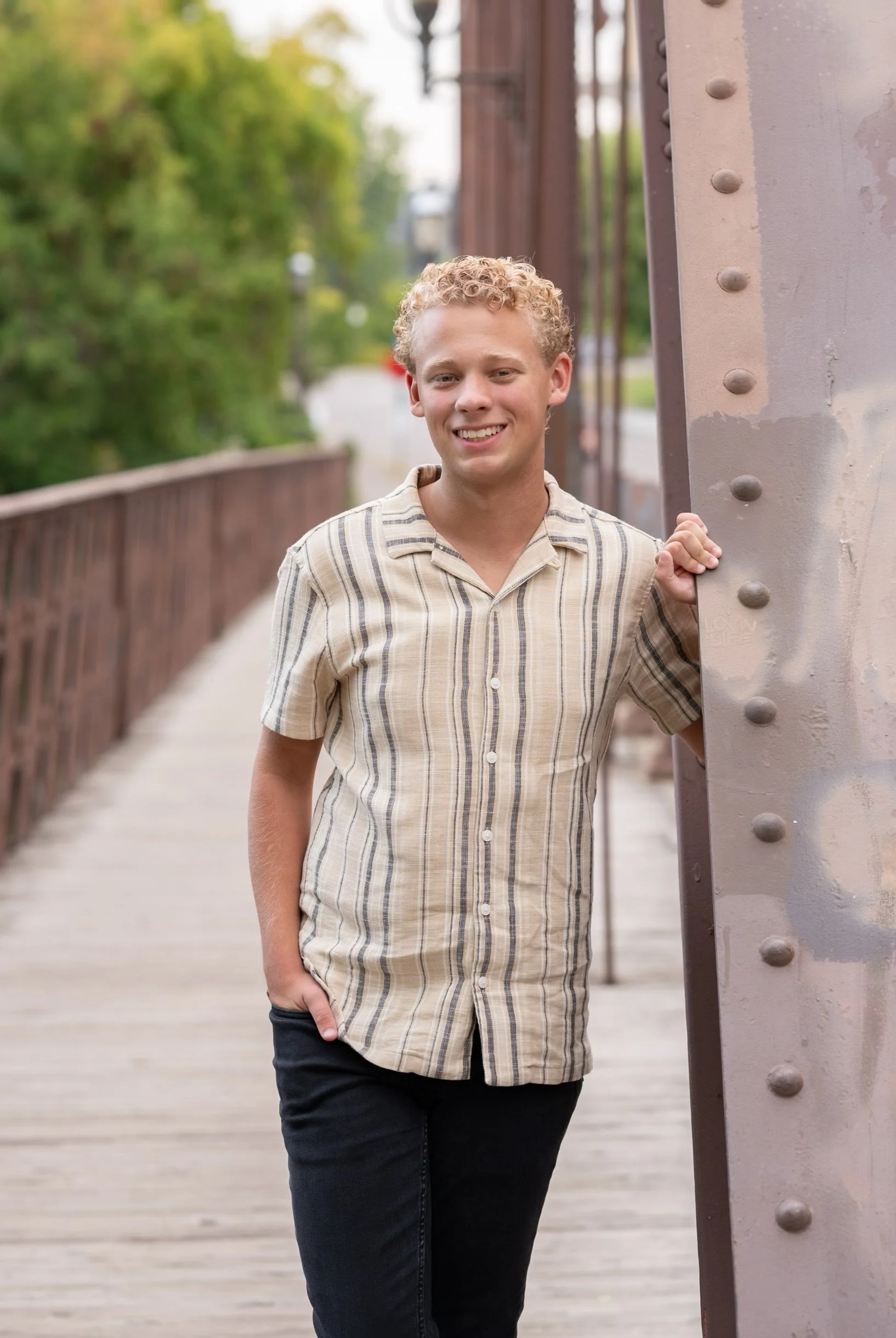 A young man with curly blonde hair and a light tan shirt with vertical stripes, smiling and leaning against a large metal bridge pillar on a wooden bridge with green trees in the background.
