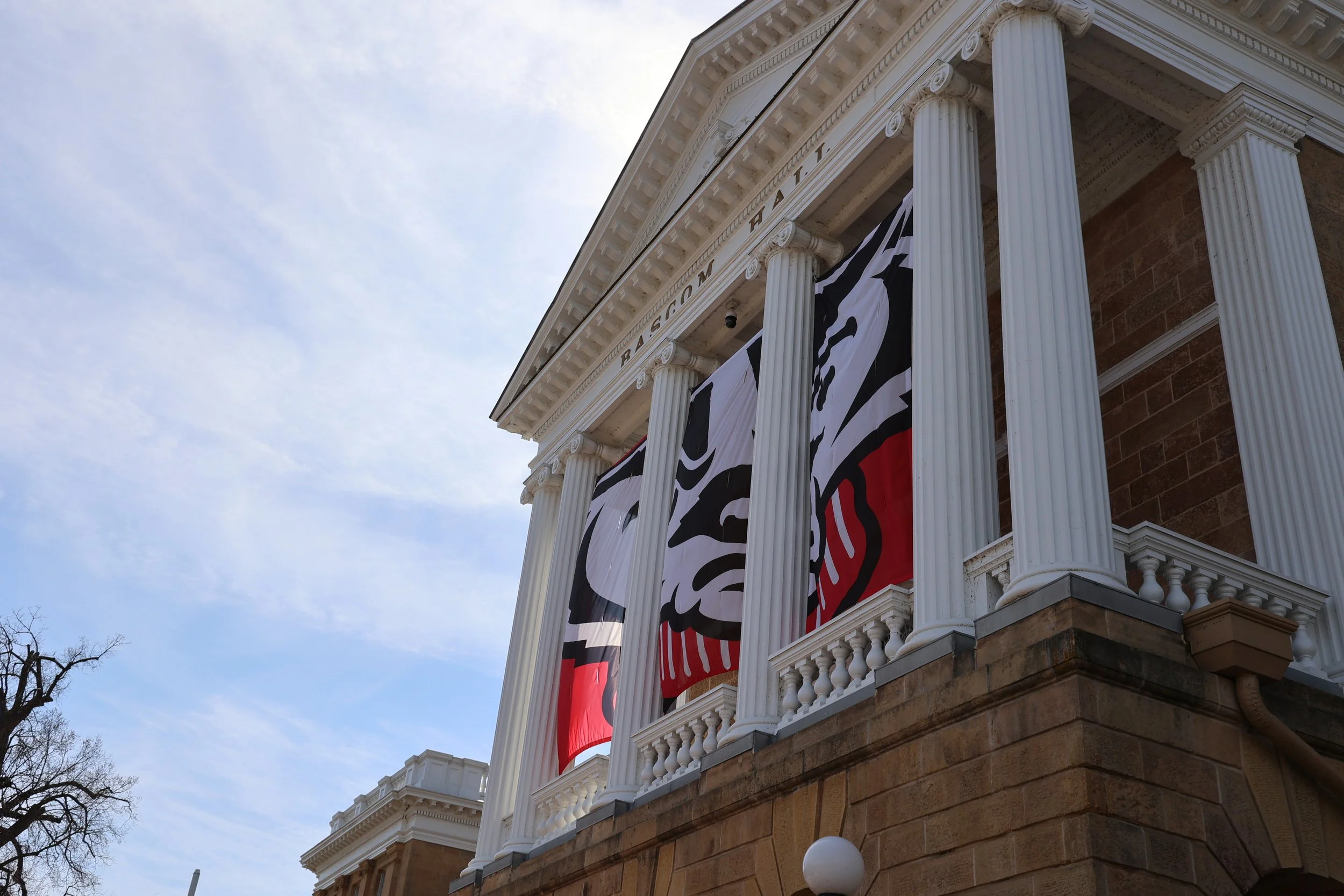 A historical building with white columns and a stone facade, flying black and white flags, under a cloudy sky.