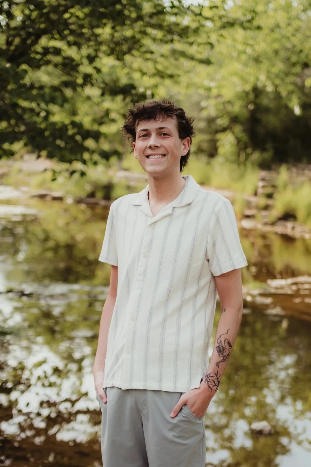 Young man with dark, curly hair and a tattoo on his forearm standing outdoors near a body of water with green trees in the background, smiling at the camera.