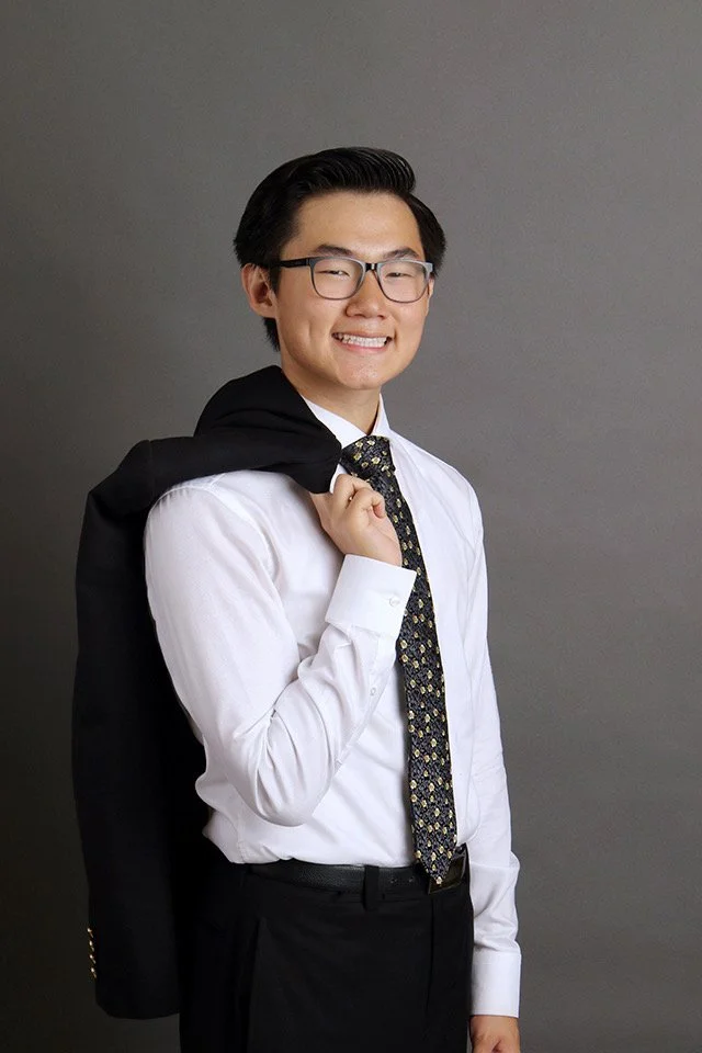 A young man with glasses, wearing a white dress shirt and a patterned tie, holding a black blazer over his shoulder, smiling, in front of a plain gray background.