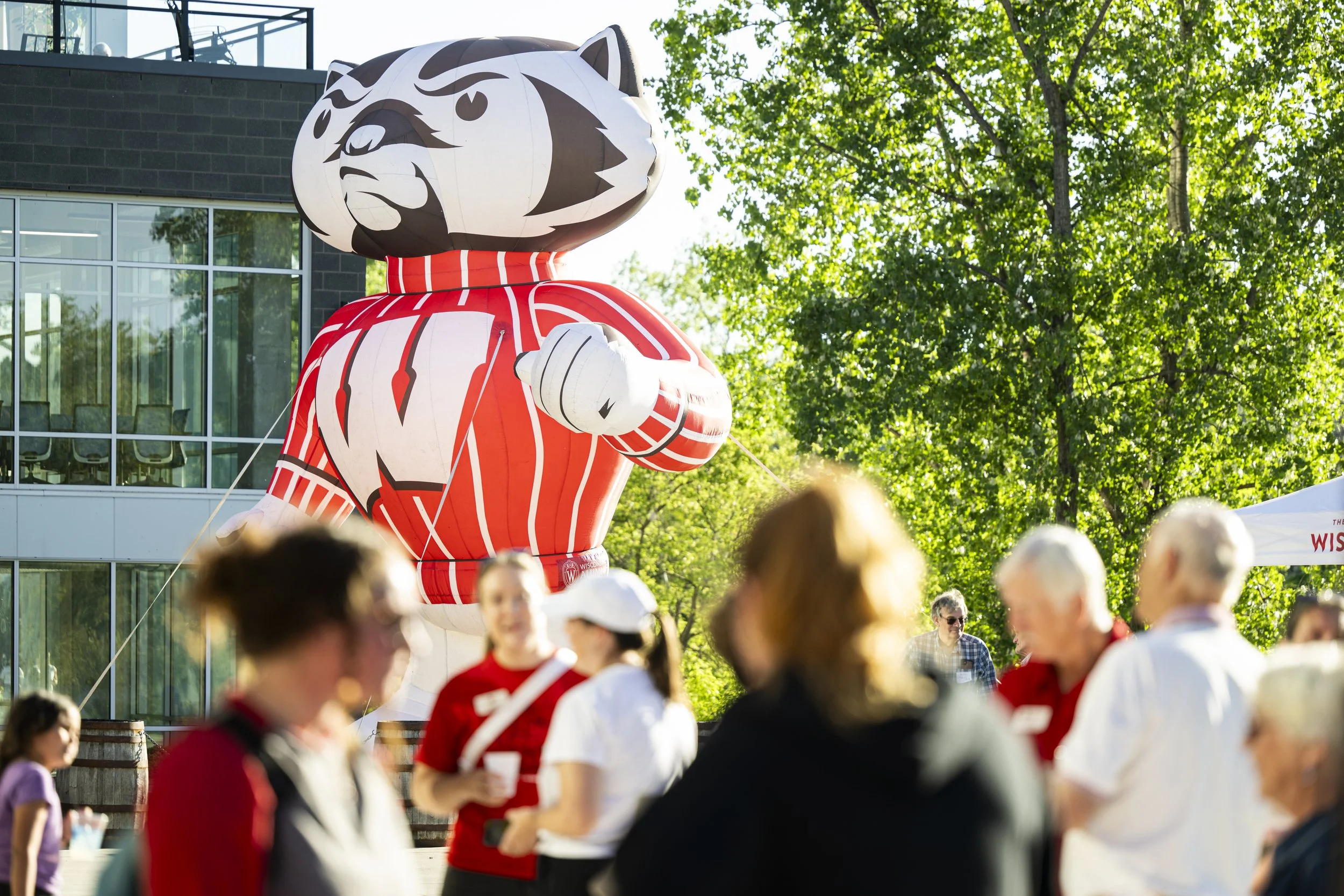A large inflatable mascot shaped like a bear wearing a red-and-white striped sweater, participating in an outdoor event with a crowd of people and green trees in the background.