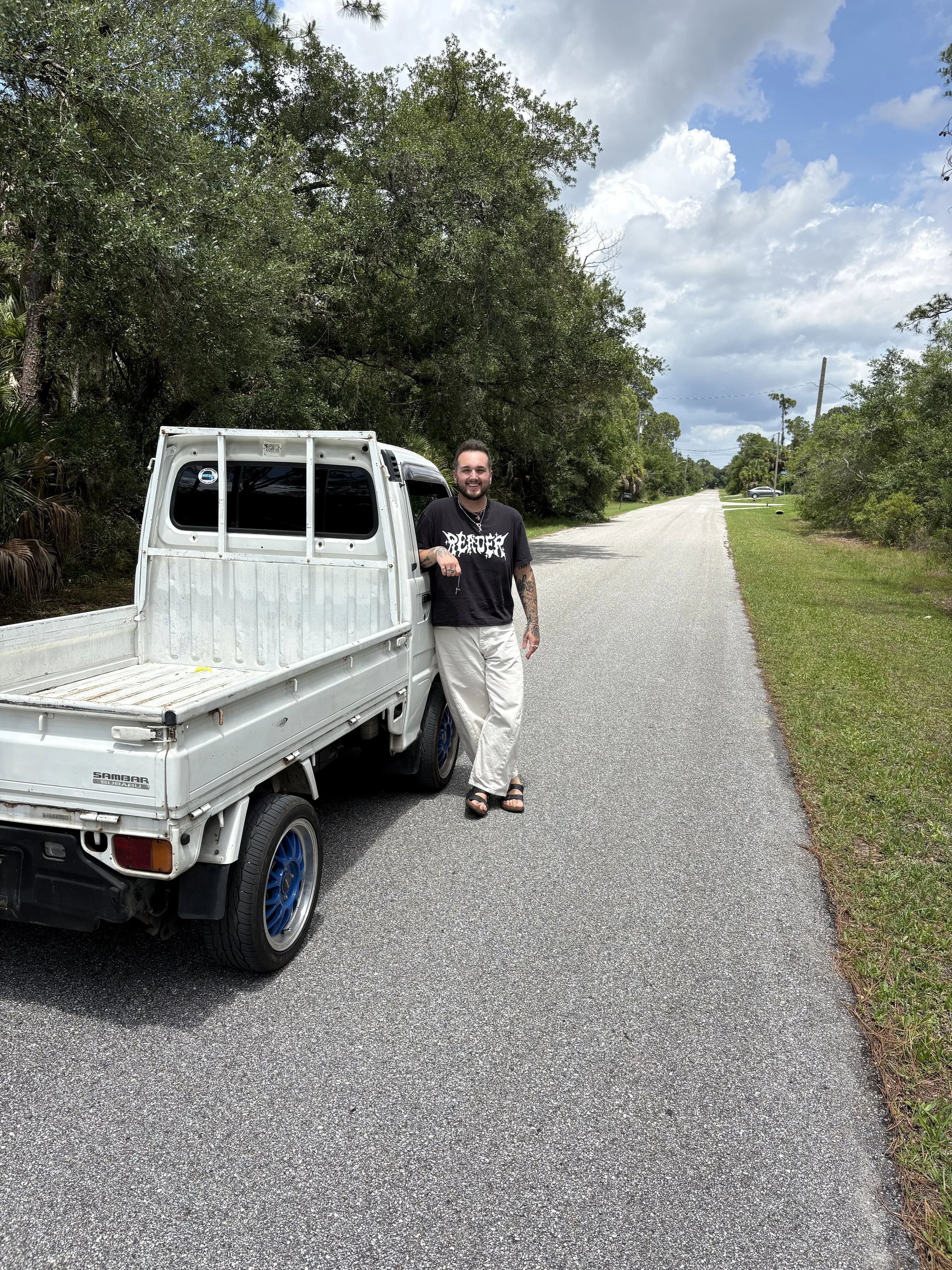 A man with tattoos and a beard smiling while leaning on the back of a small white pickup truck on a residential street with trees and clouds in the sky.