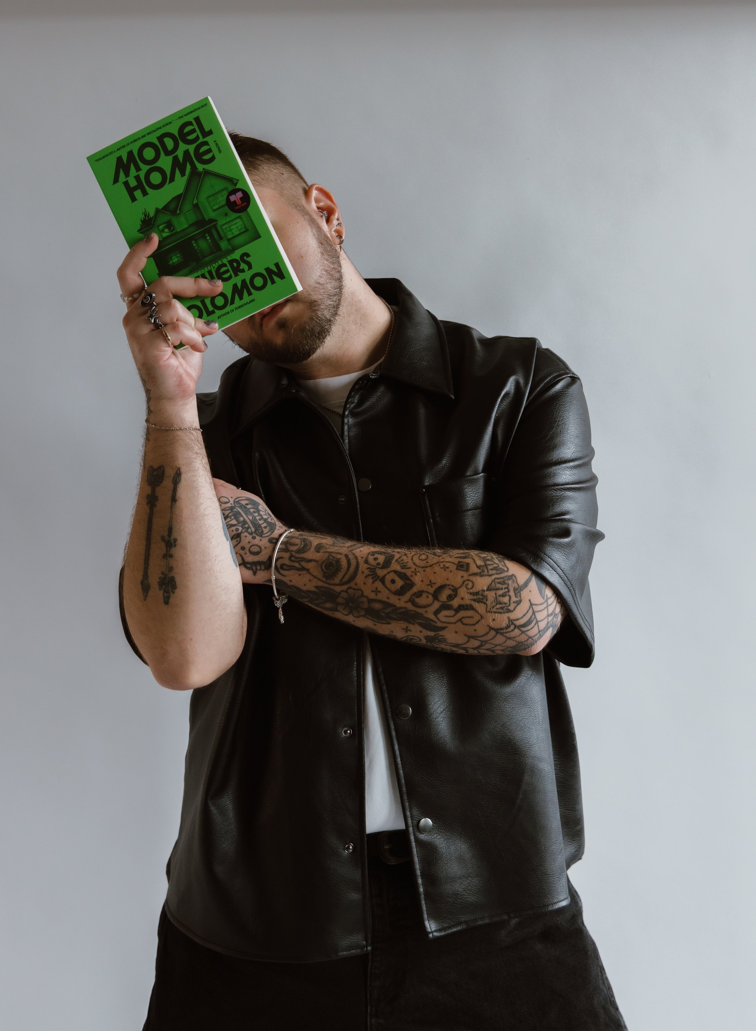 A person with tattoos on their arm, wearing a black leather jacket over a white shirt, holding a green magazine titled 'Model Home' in front of their face, with a plain light-colored background.