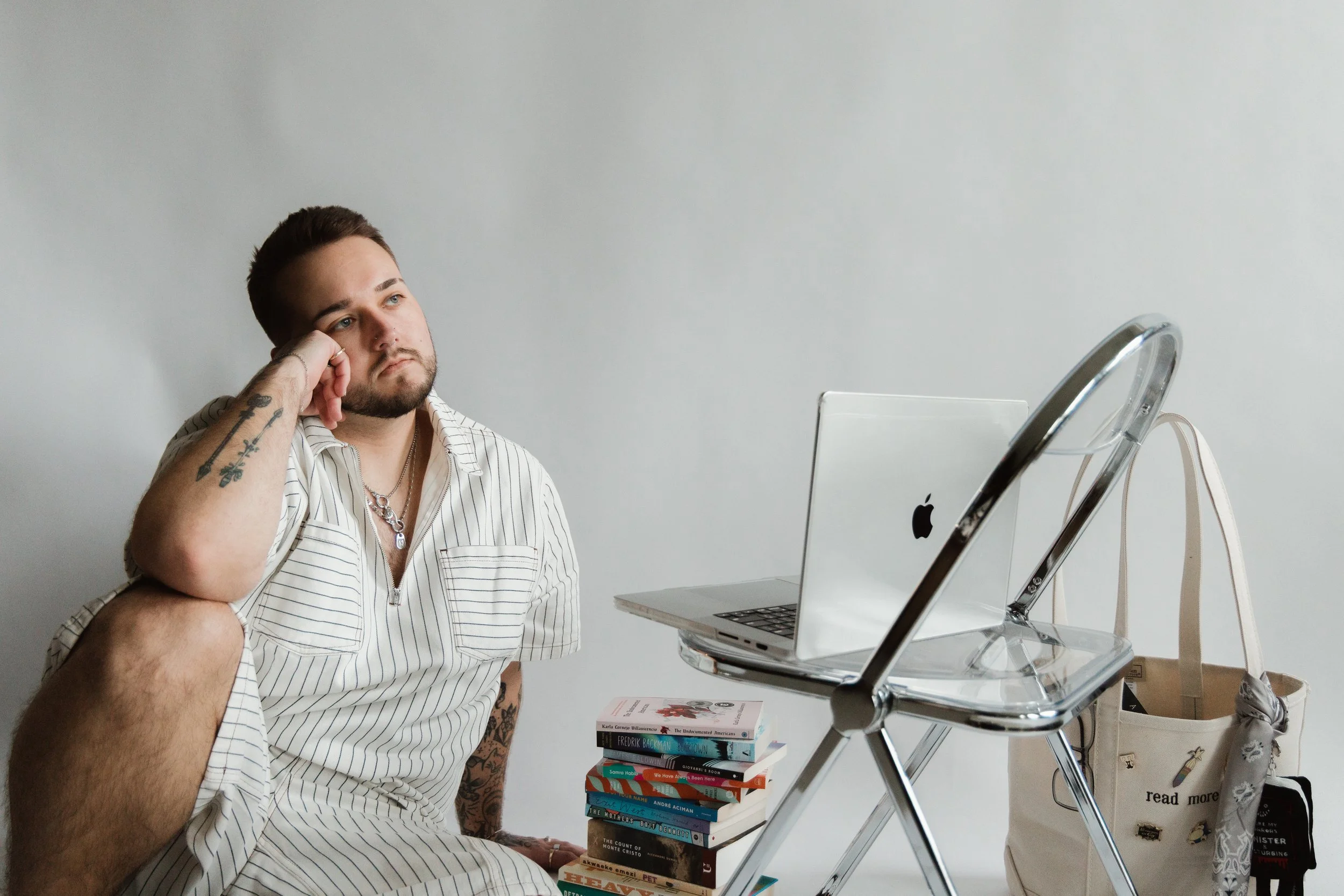 A young man with tattoos, wearing a striped shirt, sits at a table with his head resting on his hand, looking thoughtful. In front of him, there is a stack of books and an open laptop on clear stacked chairs. The background is a plain white wall.