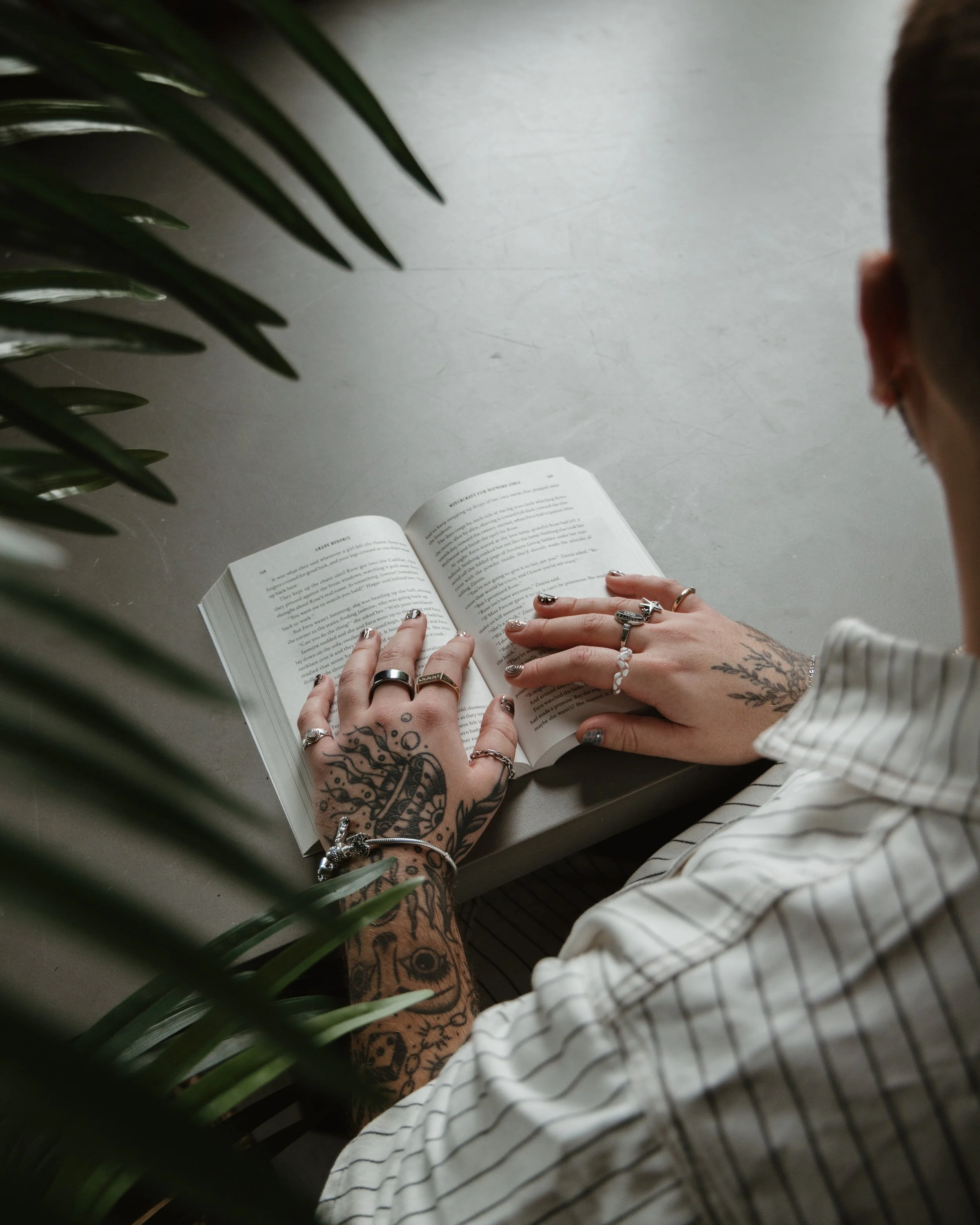 Person with tattoos on hands and wrist, wearing multiple rings and bracelets, reading a book on a gray surface with a green plant partially obscuring the view, and wearing a striped shirt.