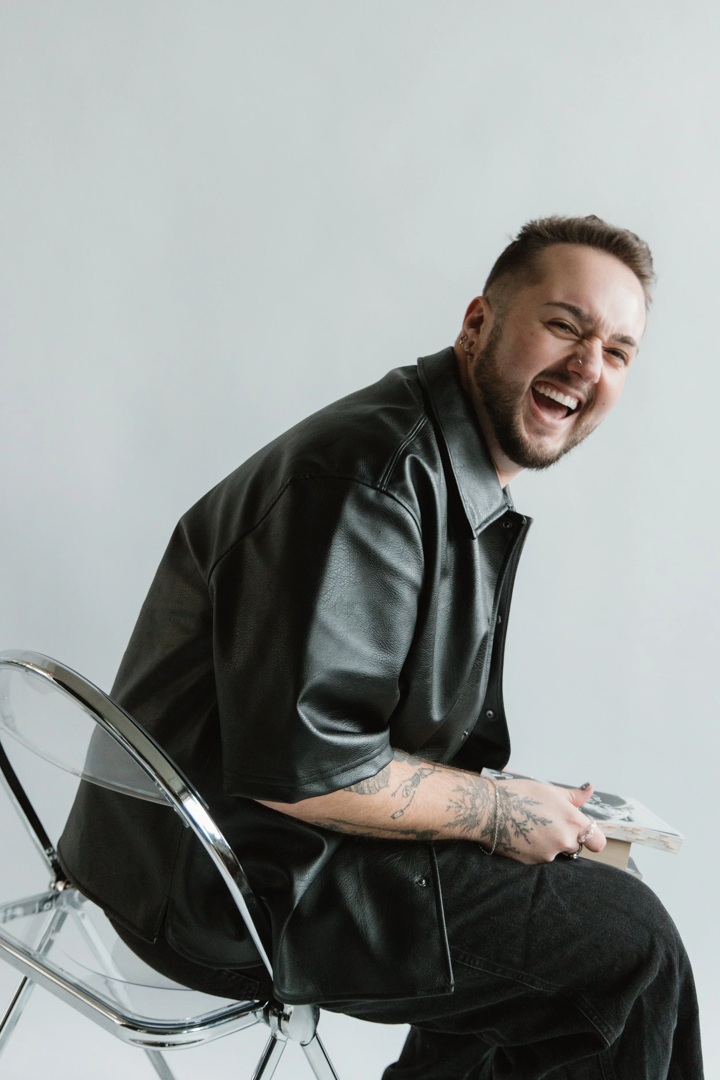 A man with a beard, tattoos, earrings, and a nose ring sitting on a clear chair, wearing a black leather jacket and black jeans, smiling and holding a magazine, against a plain white wall.