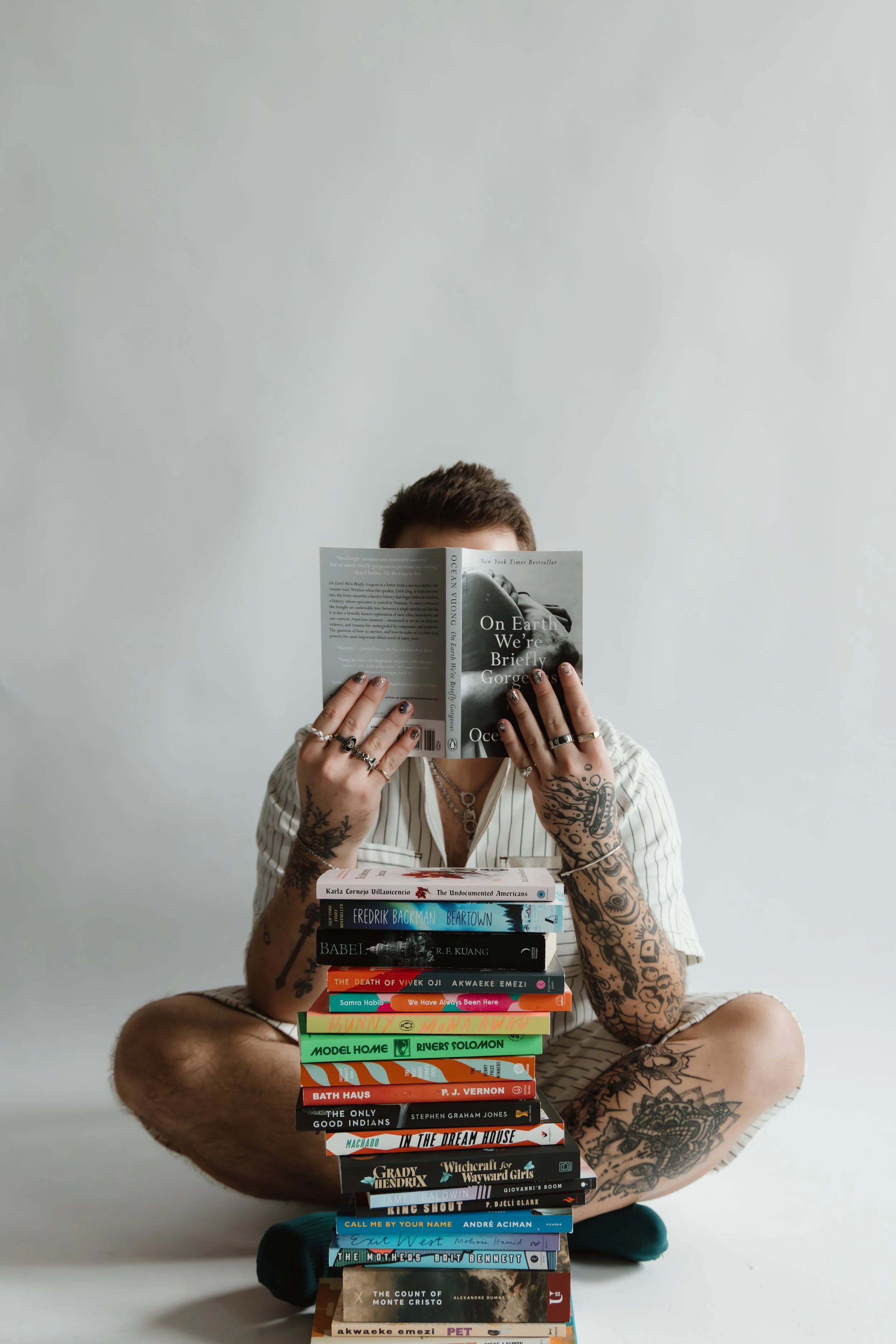 A person with tattoos on their arms and legs, wearing a striped shirt with rolled-up sleeves, sitting cross-legged, holding a book in front of their face. They are in front of a stack of multiple books and are positioned against a plain gray background.
