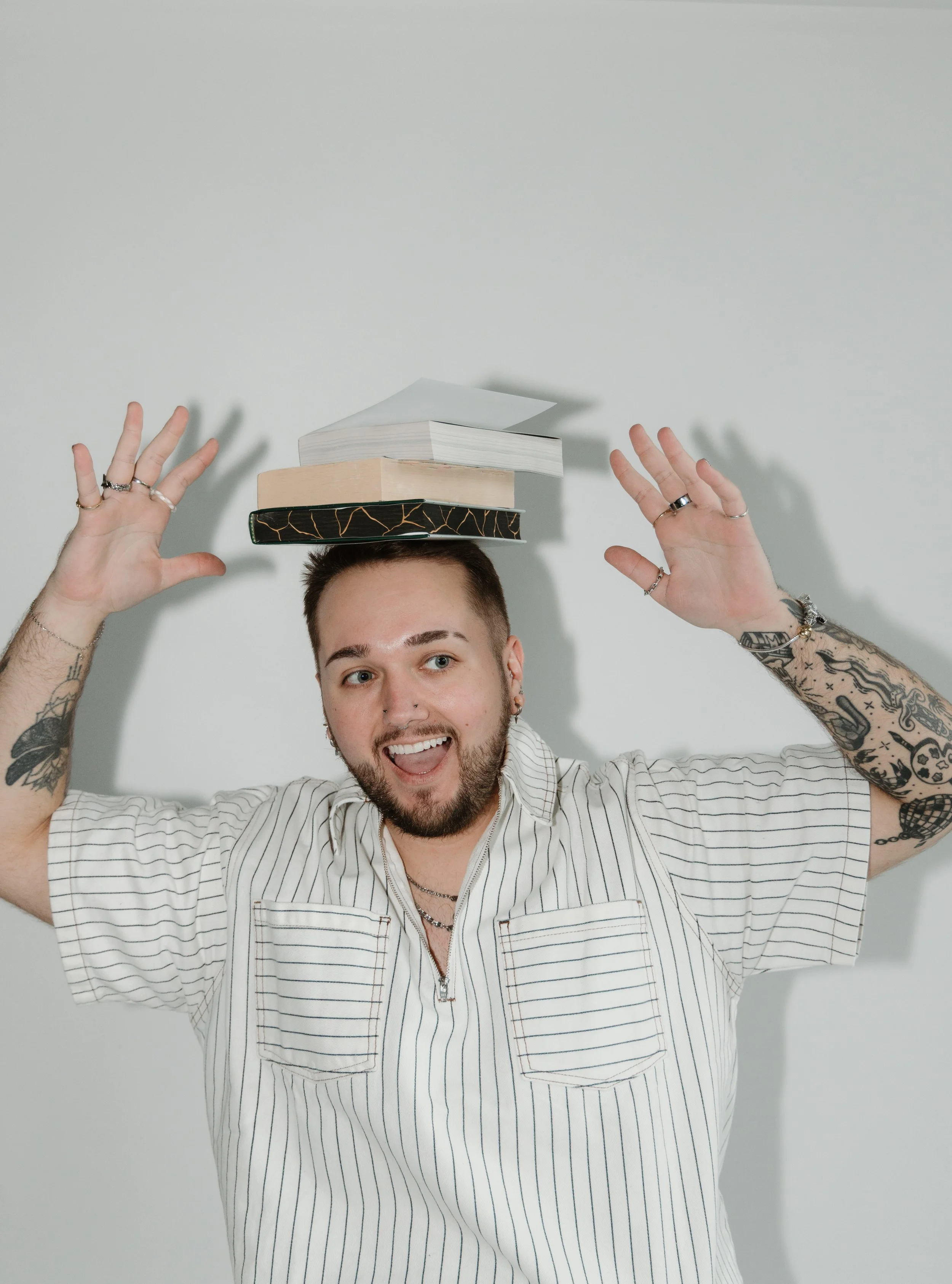 A young man with tattoos on his arms, wearing a striped shirt, is smiling and holding his hands near his head. There are three books stacked on his head, with the top book open. The background is a plain light-colored wall.