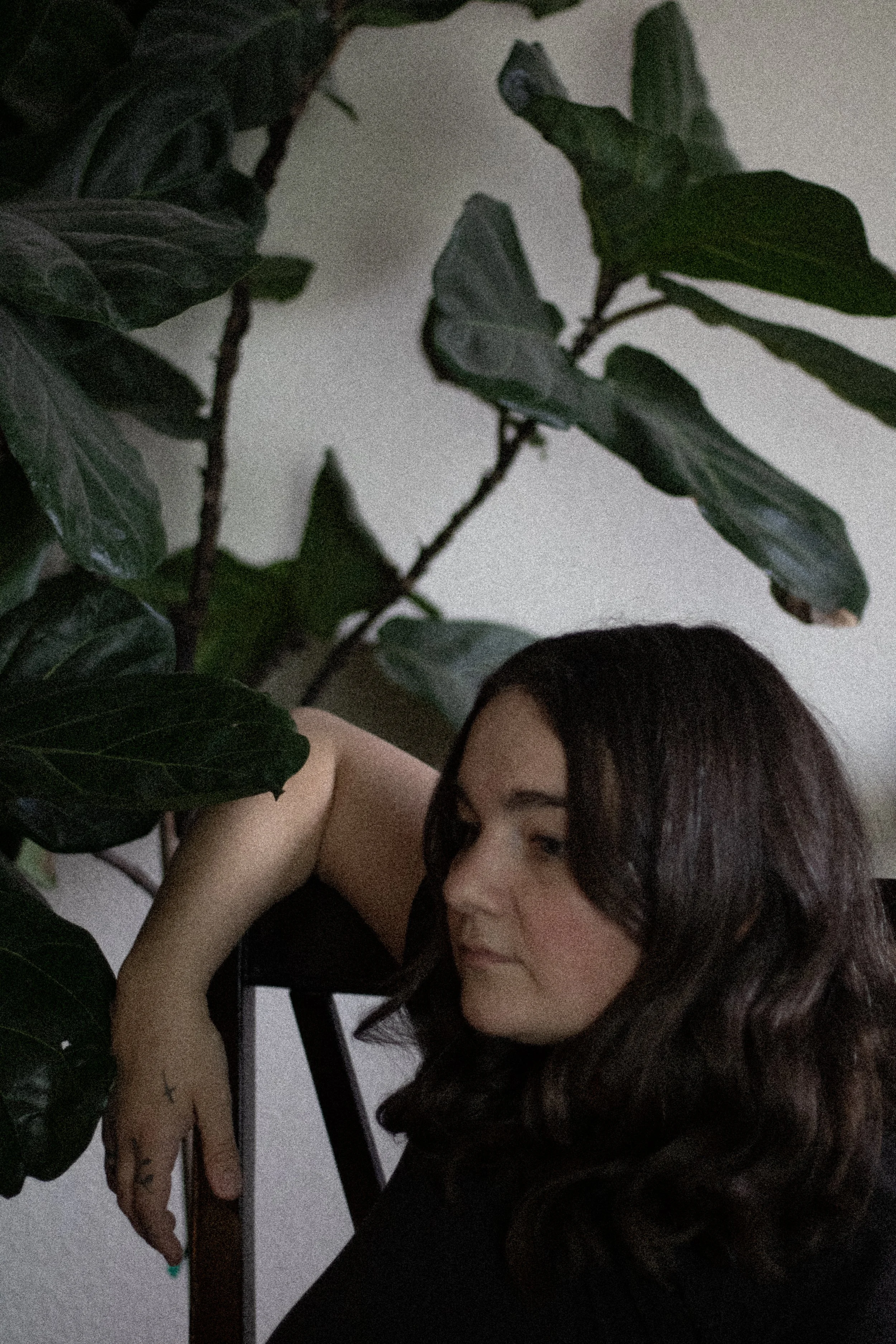 A woman with long dark hair, tattoos on her arm, and green nail polish leaning against a table next to a green leafy plant.