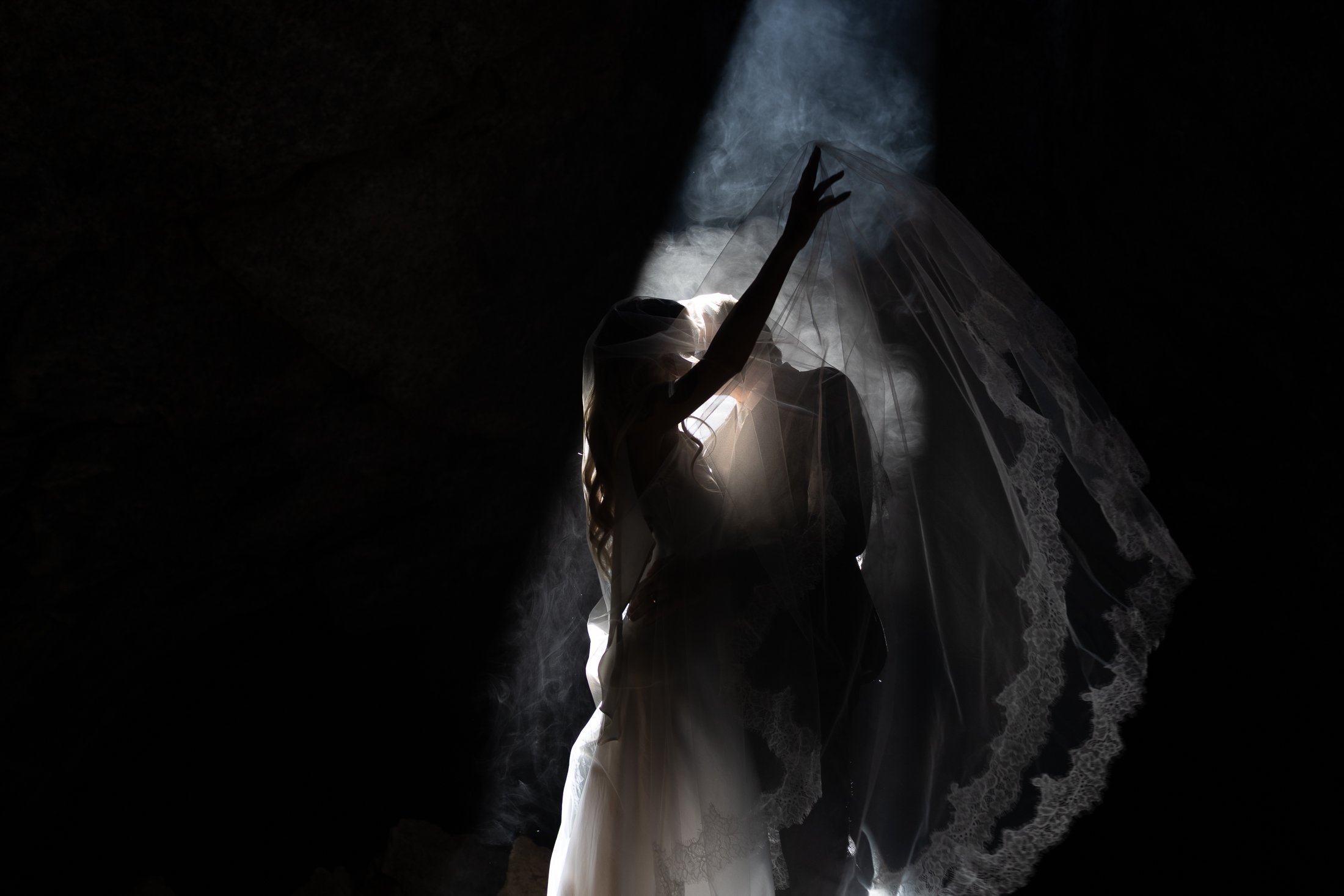 A person wearing a white dress with lace and a veil, reaching up towards a misty, illuminated point in the darkness.