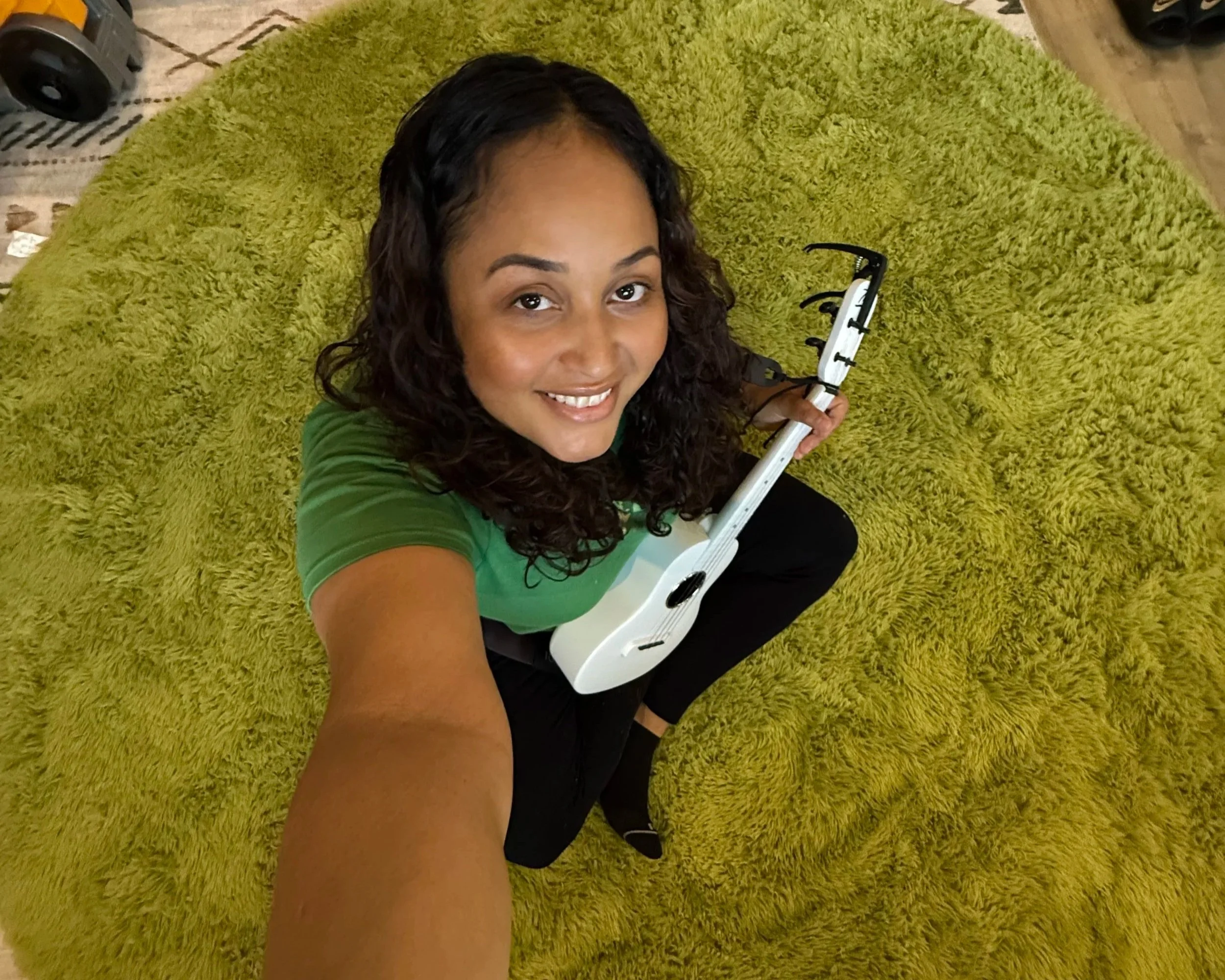 A woman with curly dark hair, smiling, taking a selfie on a green shaggy rug, holding a white guitar. She is wearing a green top and black pants, with a pair of black shoes on her feet.