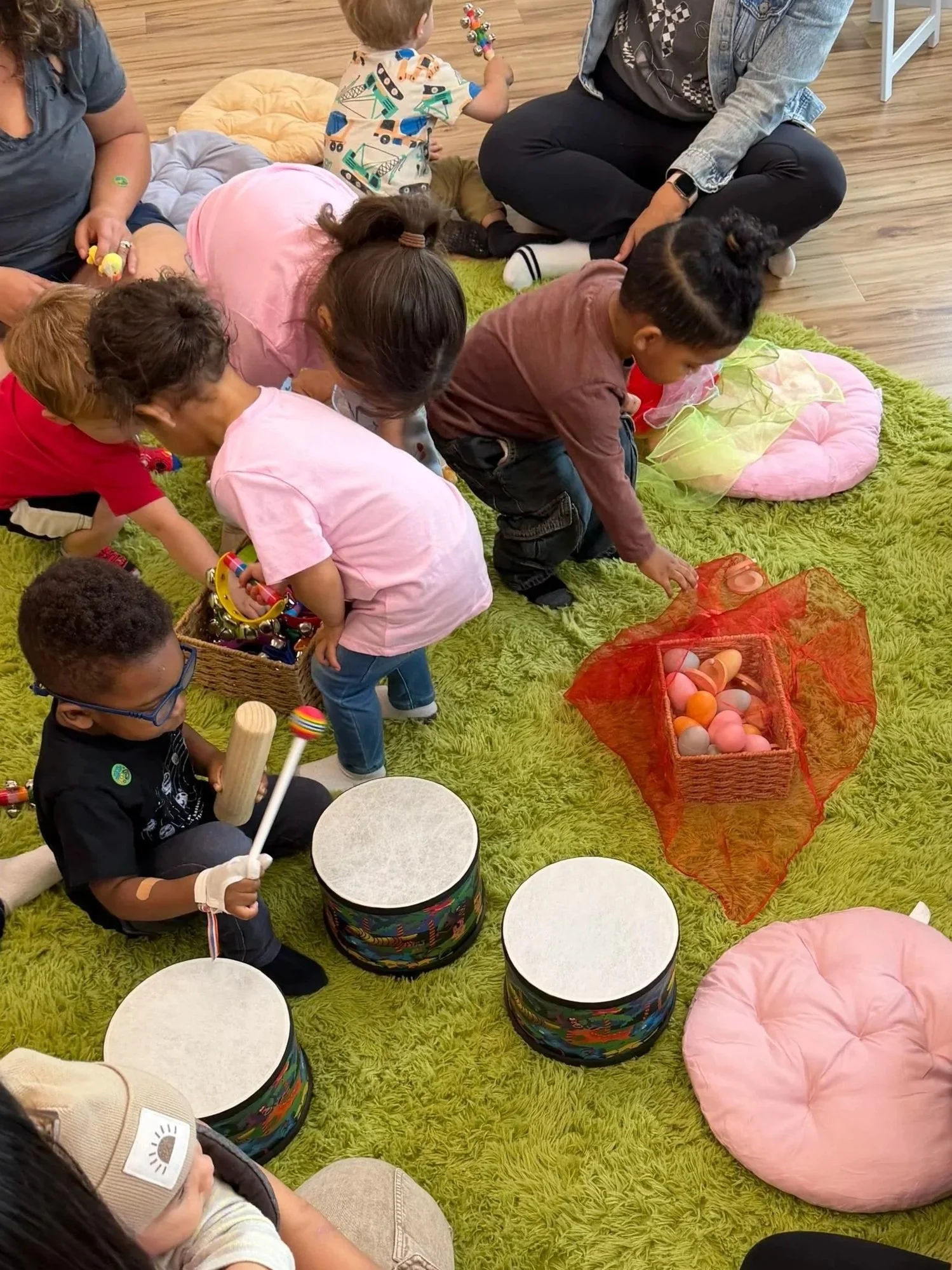 Children participating in a musical activity, sitting on a green rug, surrounded by drums, pink cushions, and colorful toys.
