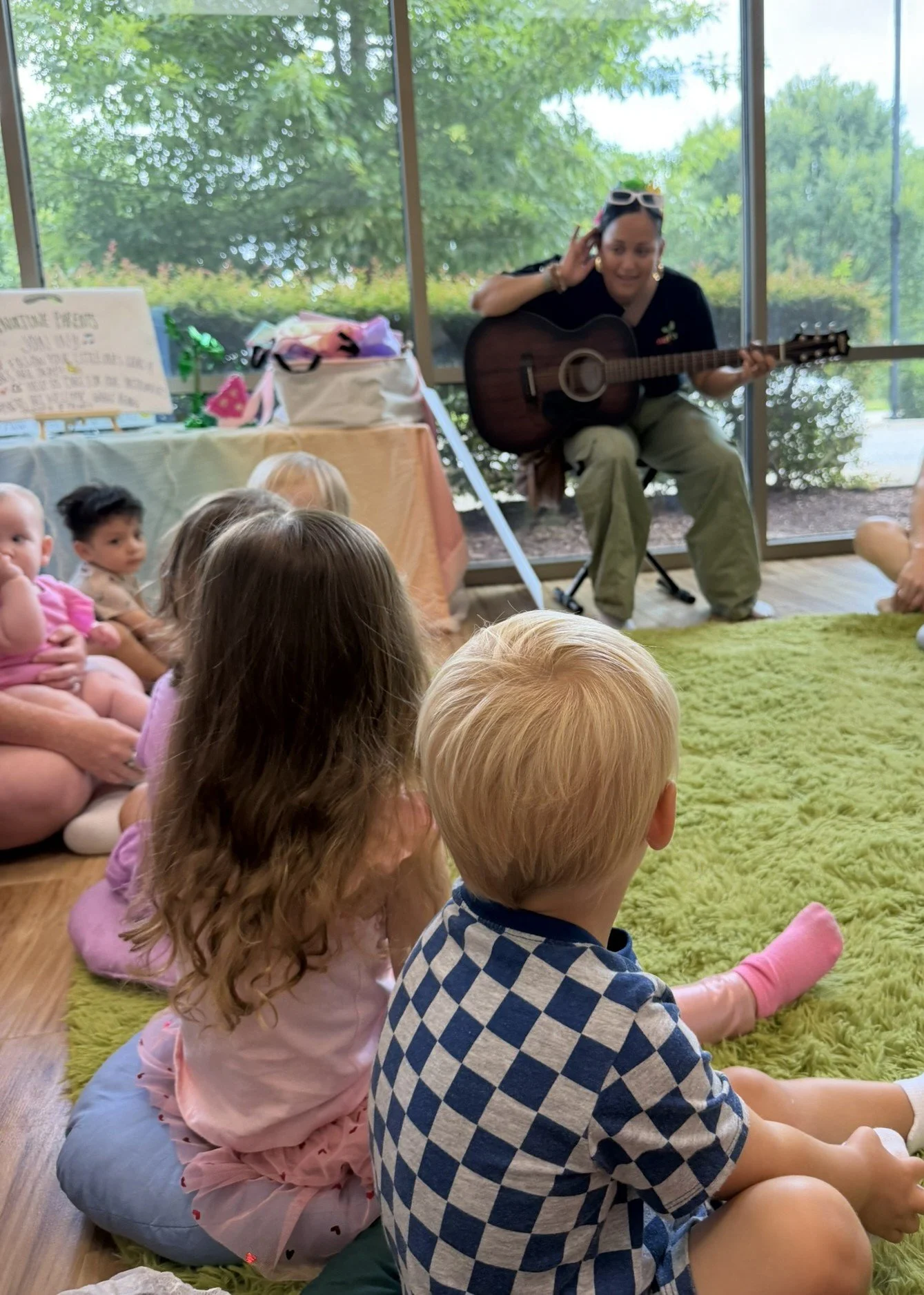 A woman playing guitar for a group of children sitting on the floor in a room with large windows and green trees outside.