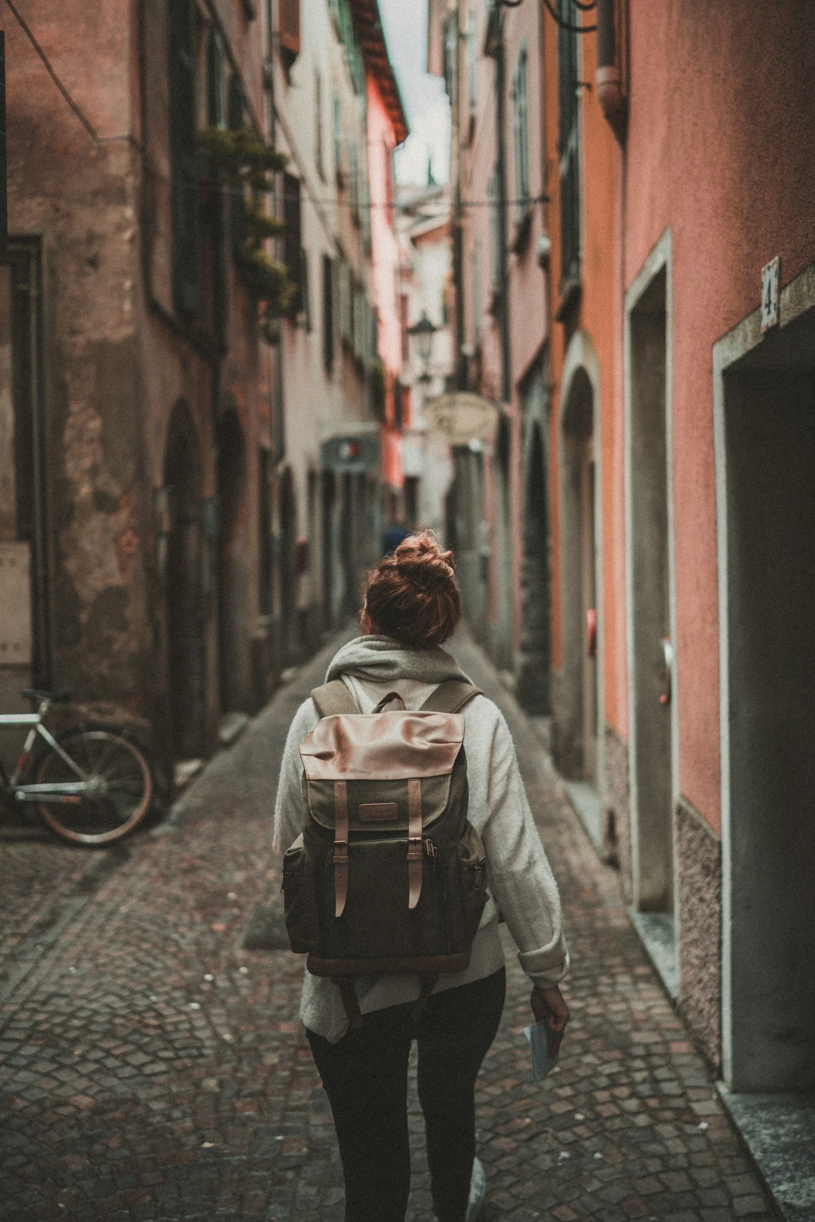 Person with bun hairstyle and backpack walking down a narrow cobblestone street lined with tall, colorful buildings.