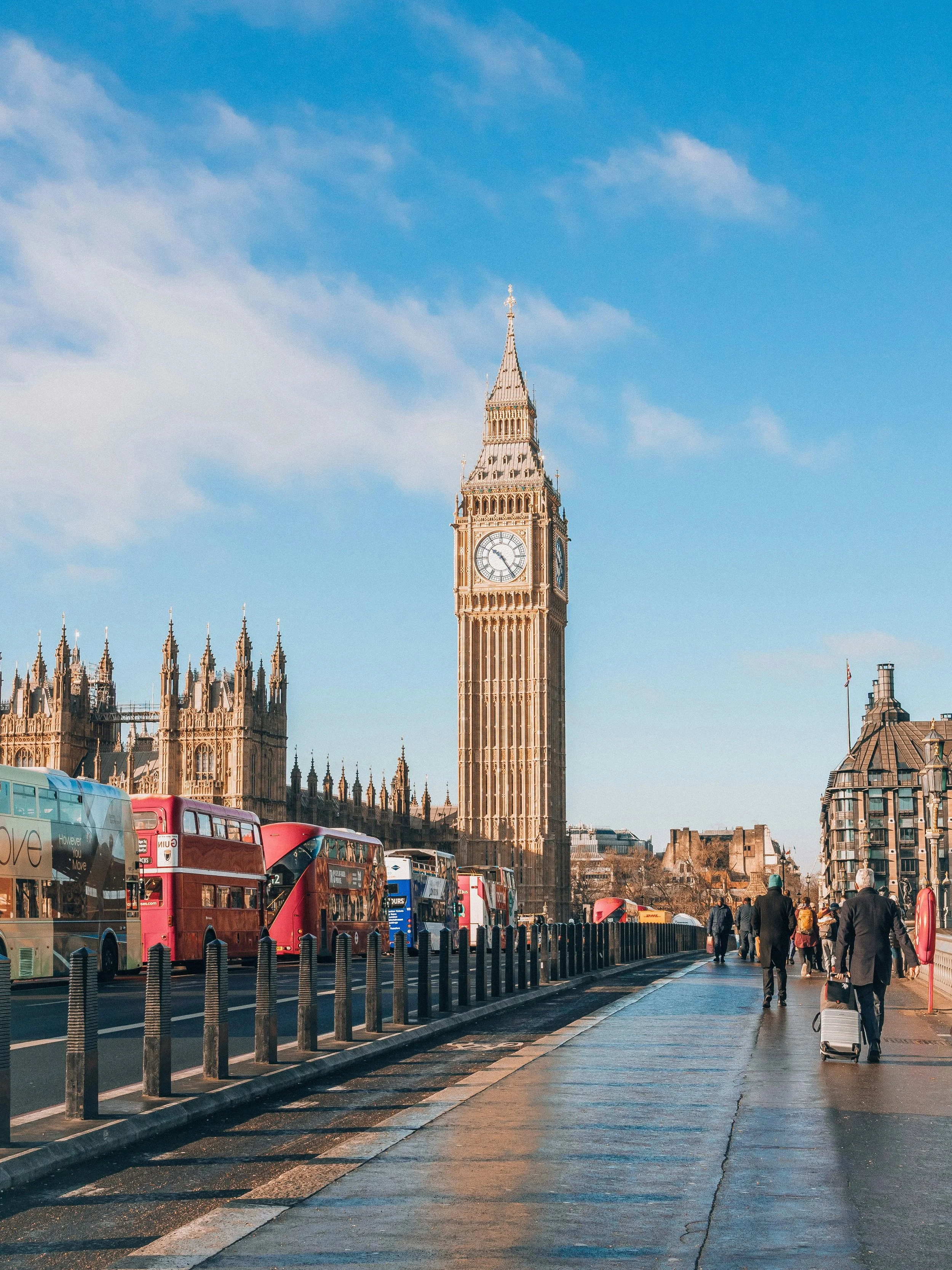 View of Big Ben clock tower in London, with a busy street and pedestrians in the foreground, and multiple buses parked along the curb.