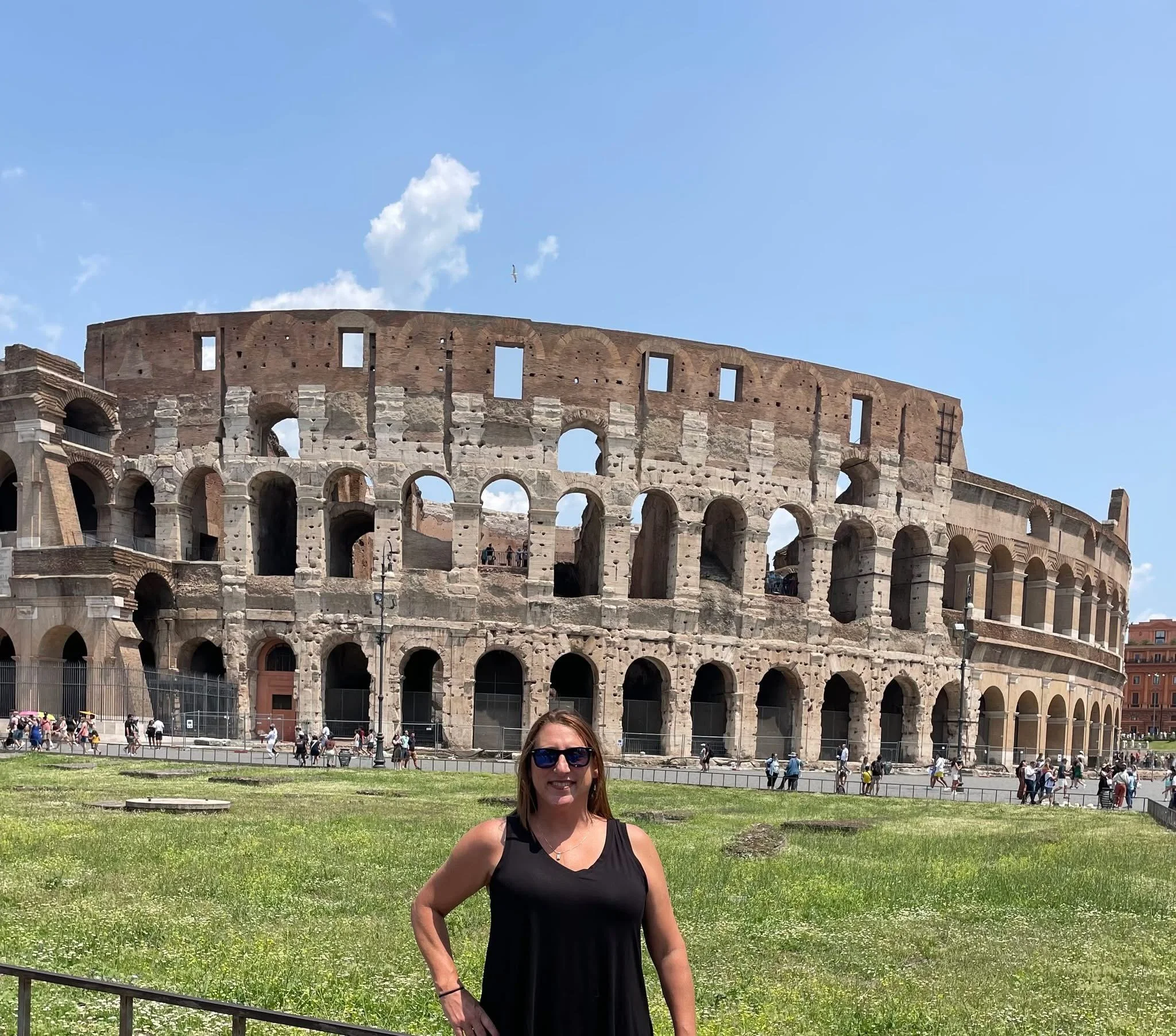 A woman with brown hair and sunglasses smiling at the camera while standing on a grassy area in front of the Colosseum in Rome, Italy during daytime.