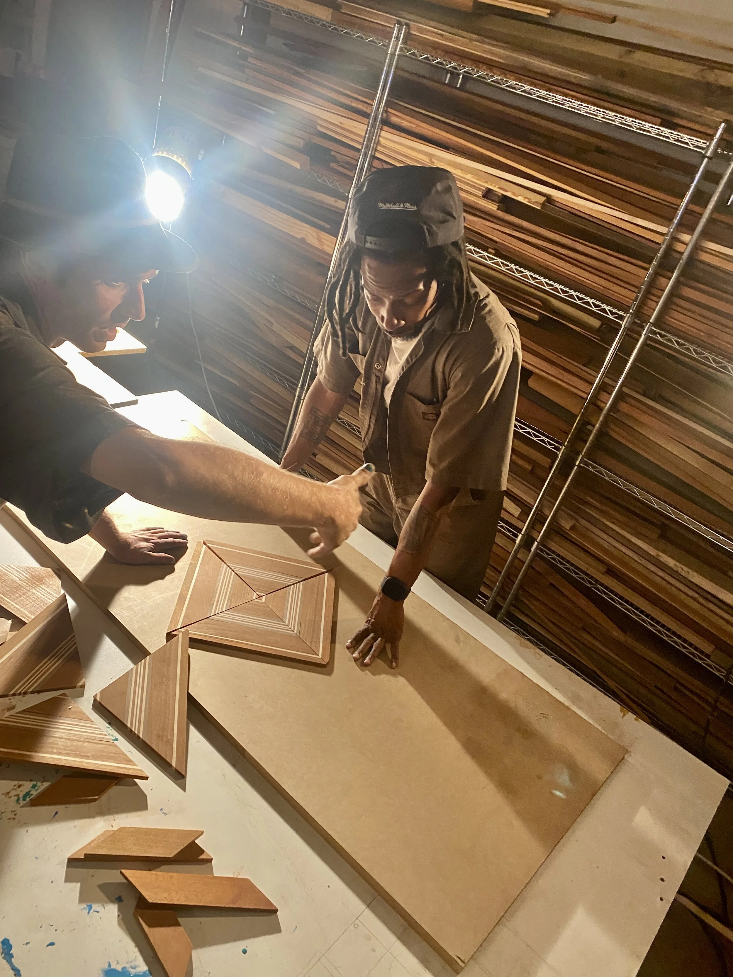Two men are assembling a geometric wooden structure on a table in a woodworking workshop. One man wears a cap and glasses with a headlamp, and the other has dreadlocks and a black cap. Shelves with wooden planks are visible in the background.