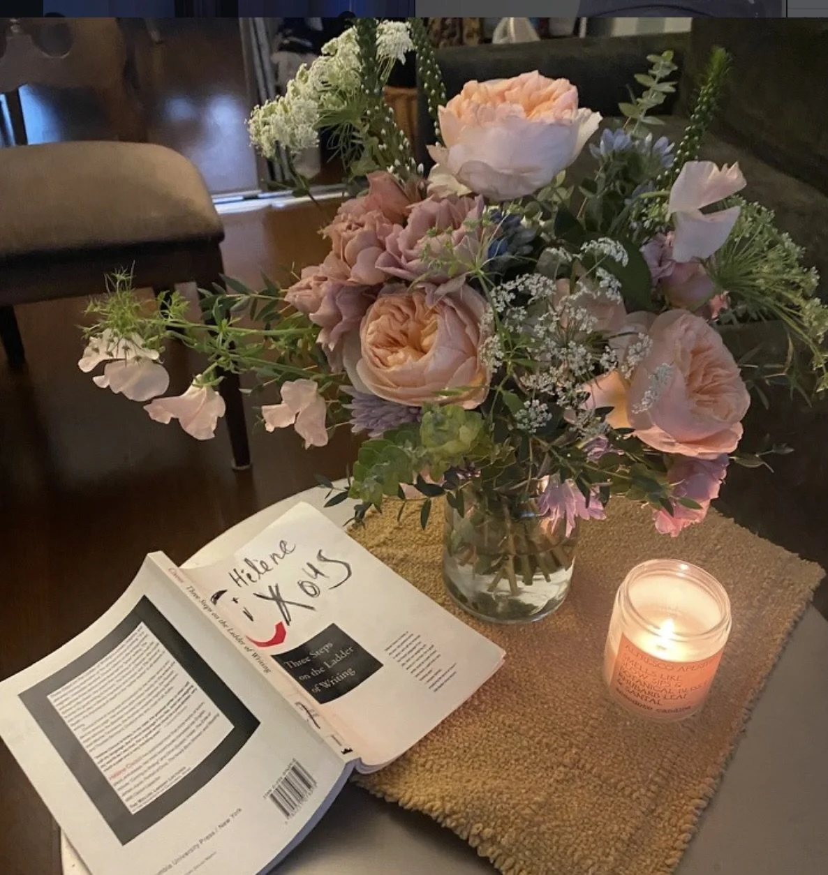 A vase of pink and white flowers, a lit candle, and an open book on a table with a woven table runner.