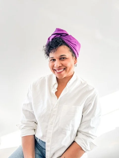 Smiling woman with short curly hair wearing a purple headwrap, white button-up shirt, and light blue jeans, standing indoors against a white background.