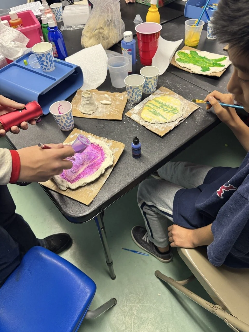 Children creating and painting on pieces of bread or dough at a table with art supplies and colorful cups.