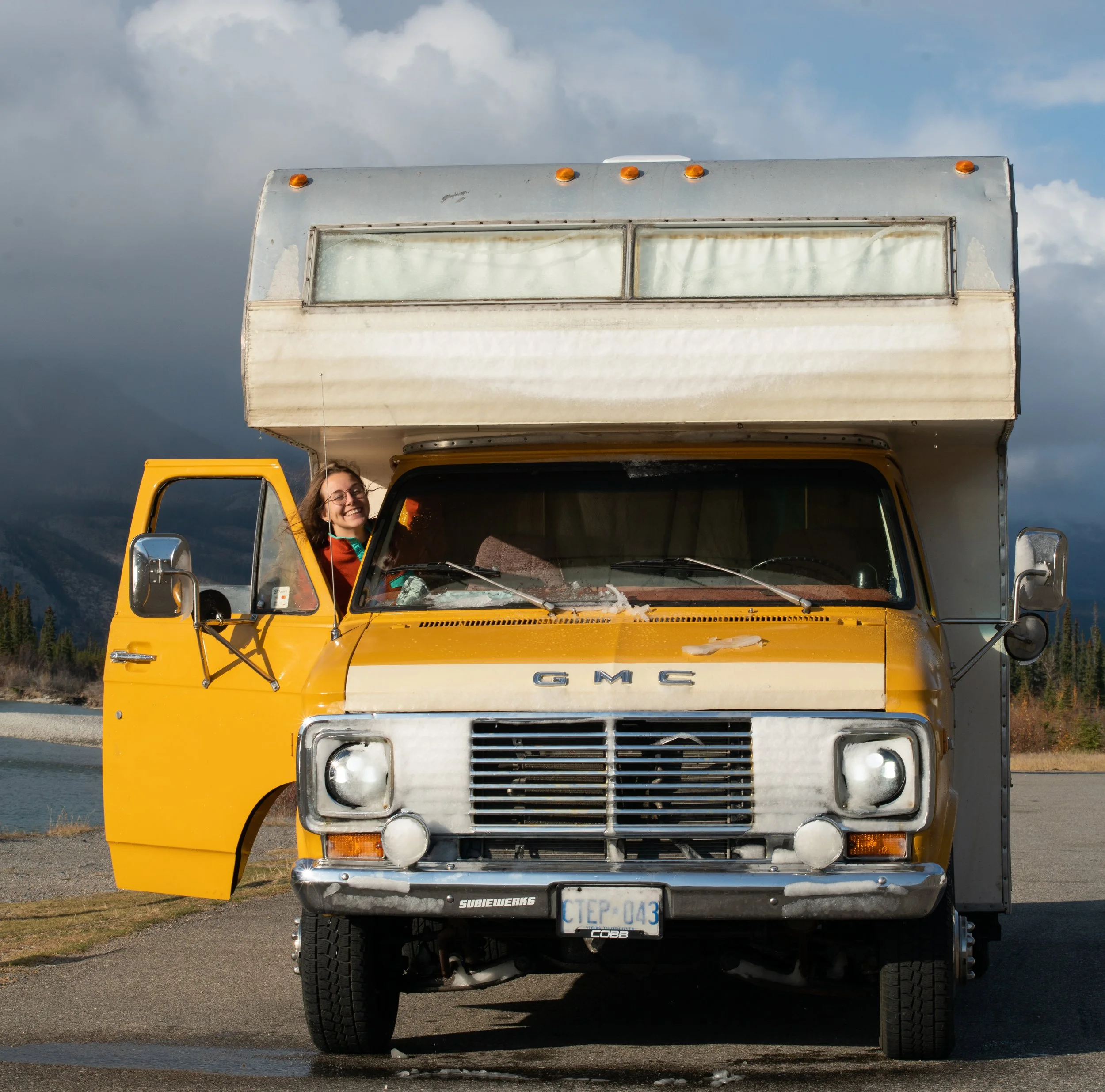 Front view of a yellow GMC motorhome with the front overhanging the drivers cabin parked outdoors under a cloudy sky, with mountains and trees in the background of Jasper AB.