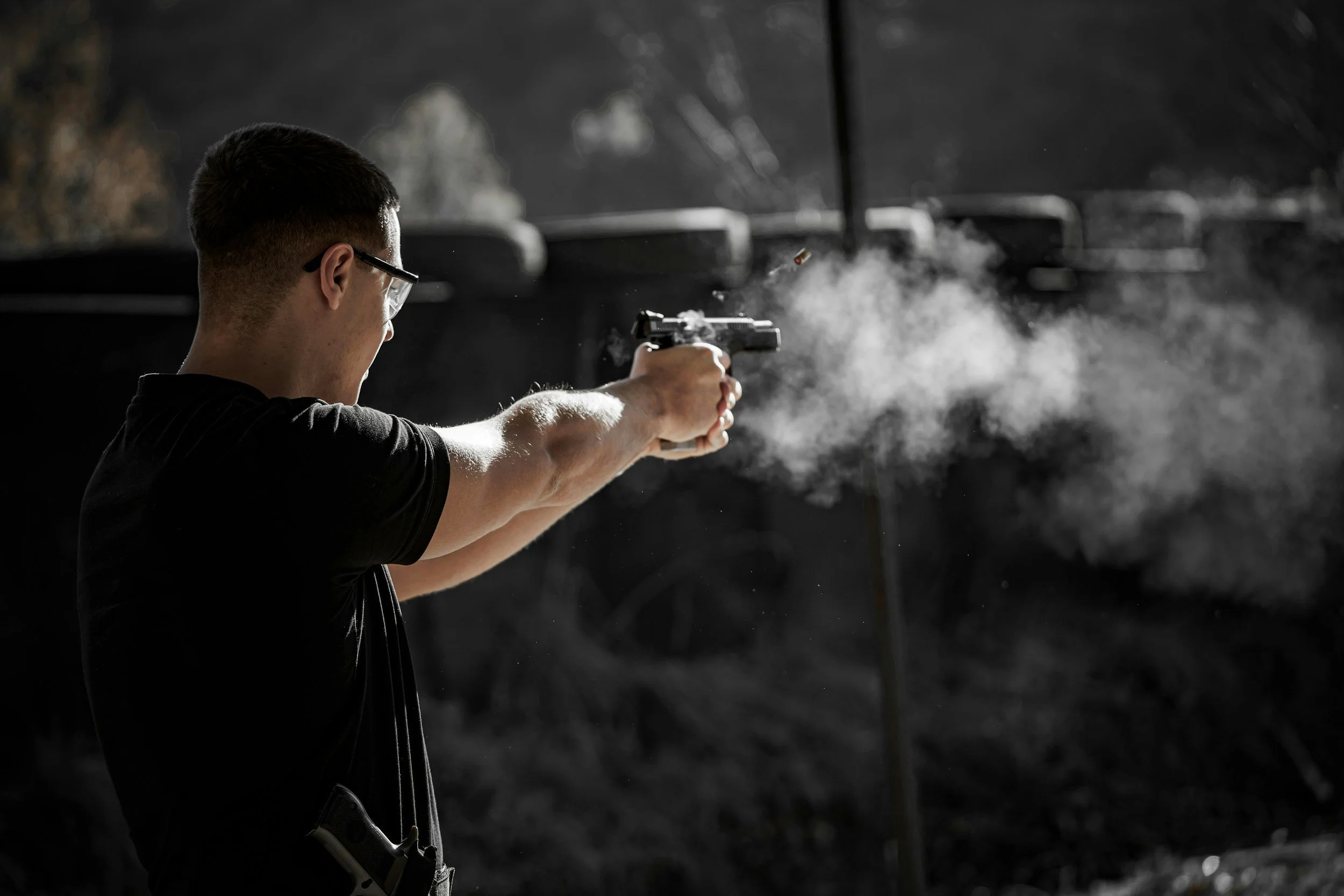A shooter at an indoor firing range lane firing a handgun showing the off gas coming out of the gun.