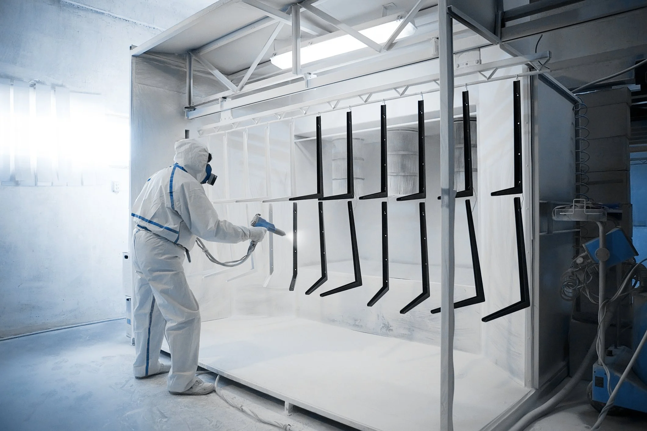 A powder coating technician in PPE spraying coating while the cylindrical exhaust filters collect powder overspray.