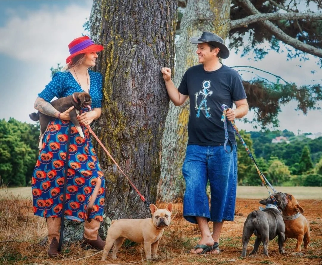 A woman holding a small brown dog and a man standing with three dogs outdoors near a large tree, with a grassy background and a cloudy sky.