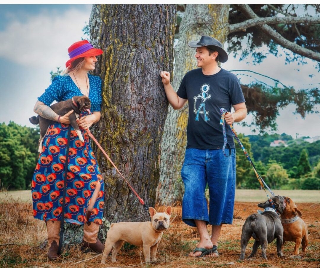 Olivia and Alistair Potter of Le Epitome Kennels standing outdoors near a tree at Auckland Botanic gardens with coloured French Bulldogs Eden, Pluto, and Mickey, alongside Tim Tam, our chocolate and tan Pug stud dog.