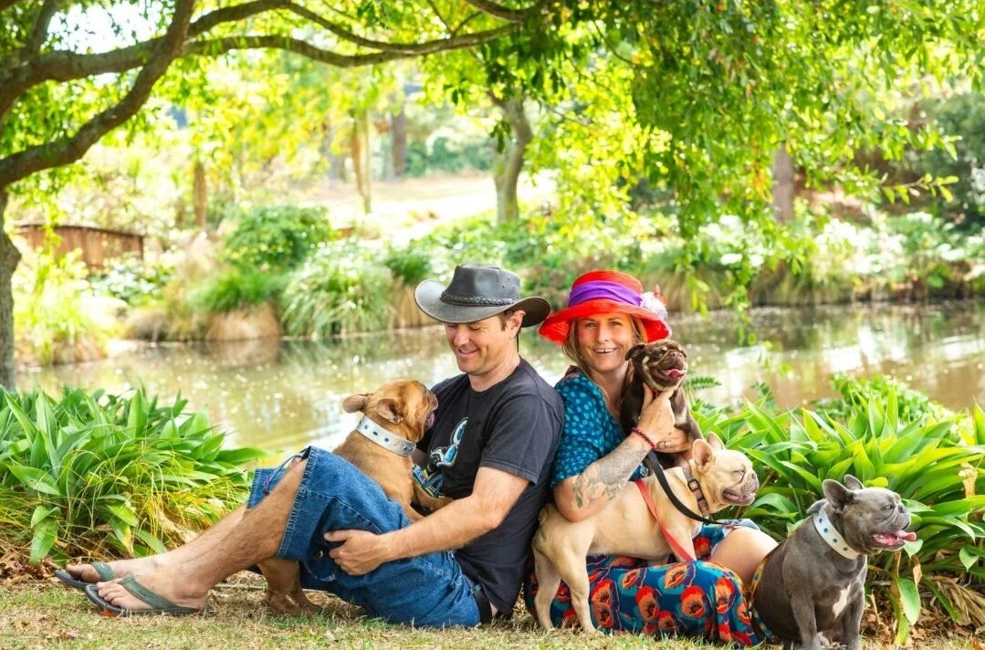 Alistair and Olivia Potter of Le Epitome Kennels in Waikato, New Zealand, sitting back to back outdoors beside a pond with their coloured French Bulldogs, and Tim Tam their Pug stud dog.