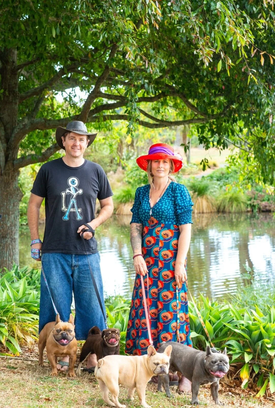 Alistair and Olivia Potter of Le Epitome Kennels in Waikato, New Zealand, standing outdoors by a pond at Auckland Botanic Gardens with three coloured French Bulldogs and a coloured Pug from their breeding programme.