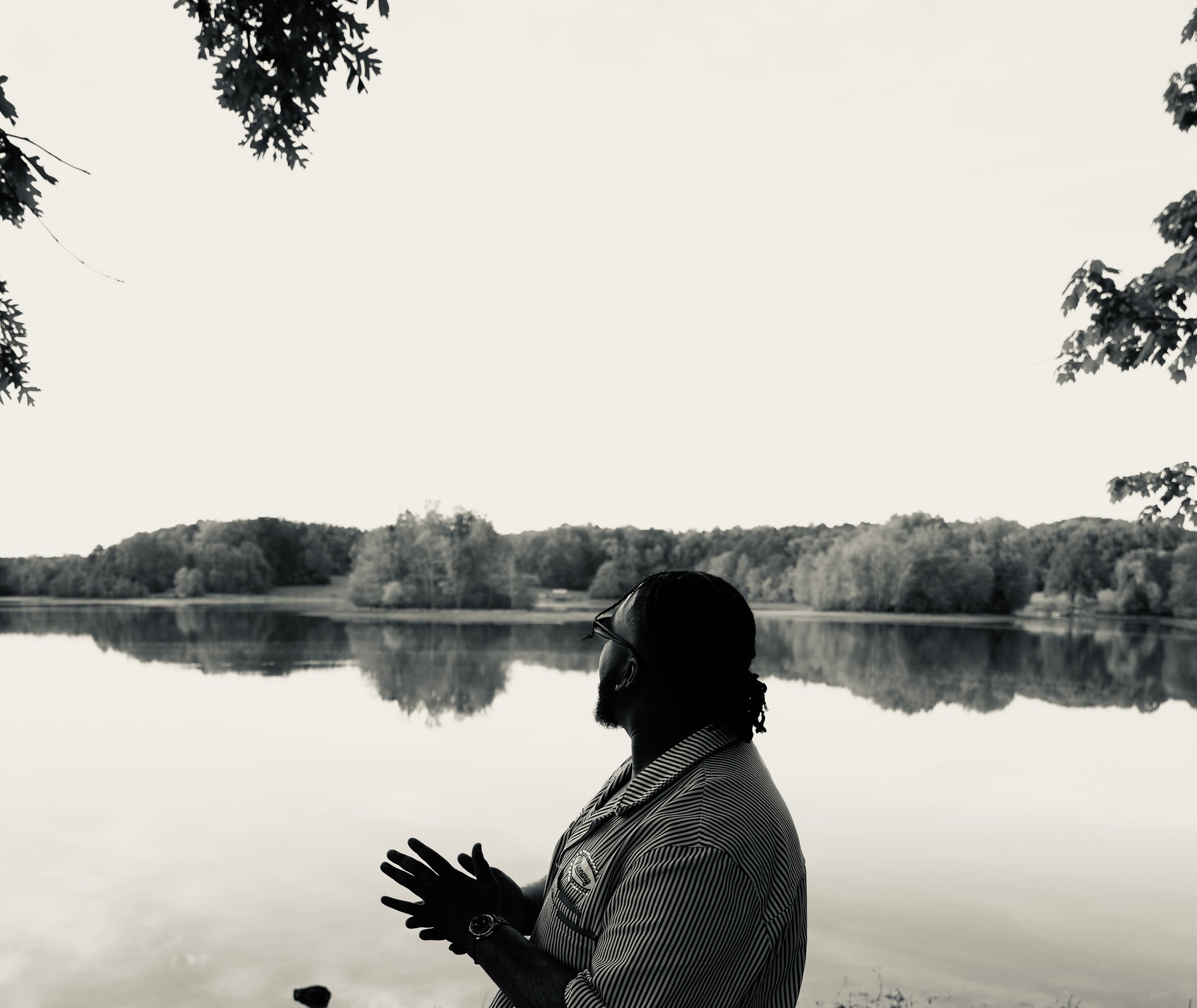 A man with braided hair wearing a striped shirt standing by a lake with trees and hills in the background, reflected in the water, as he looks to the side and gestures with his hand.