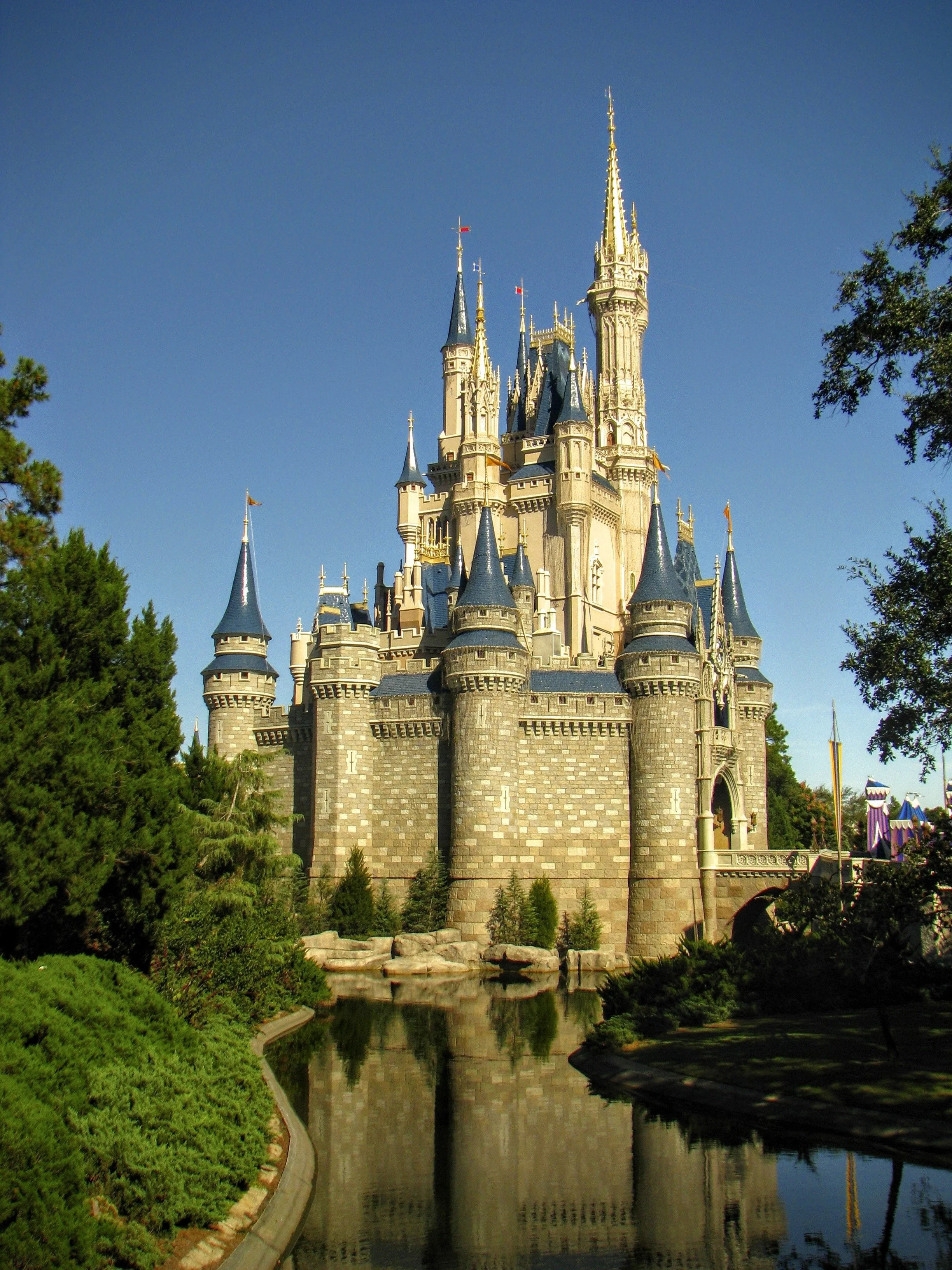 A castle with tall spires and towers, surrounded by trees and a reflecting pool in the foreground, set against a clear blue sky.