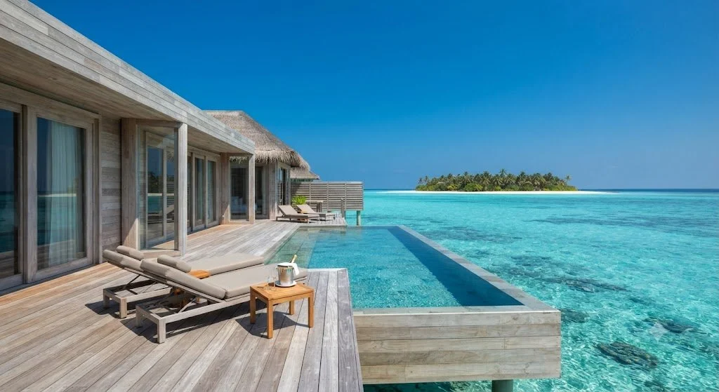 Luxury overwater bungalow with a deck, sun loungers, and an infinity pool overlooking the turquoise ocean and a small island in the distance.