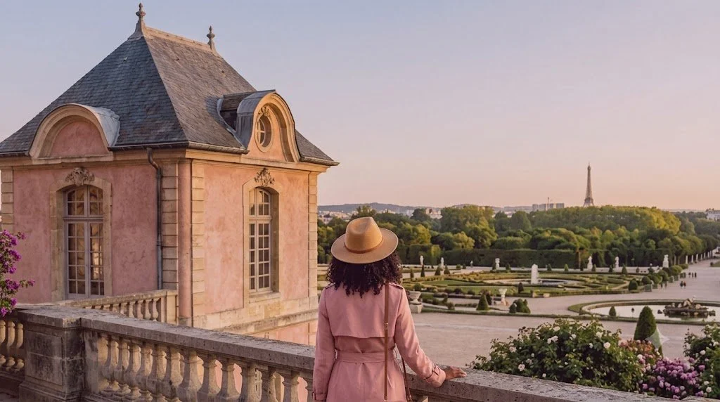 Woman with curly hair wearing a pink coat and hat looking over the gardens of the Tuileries in Paris, with the Eiffel Tower in the distance during sunset.