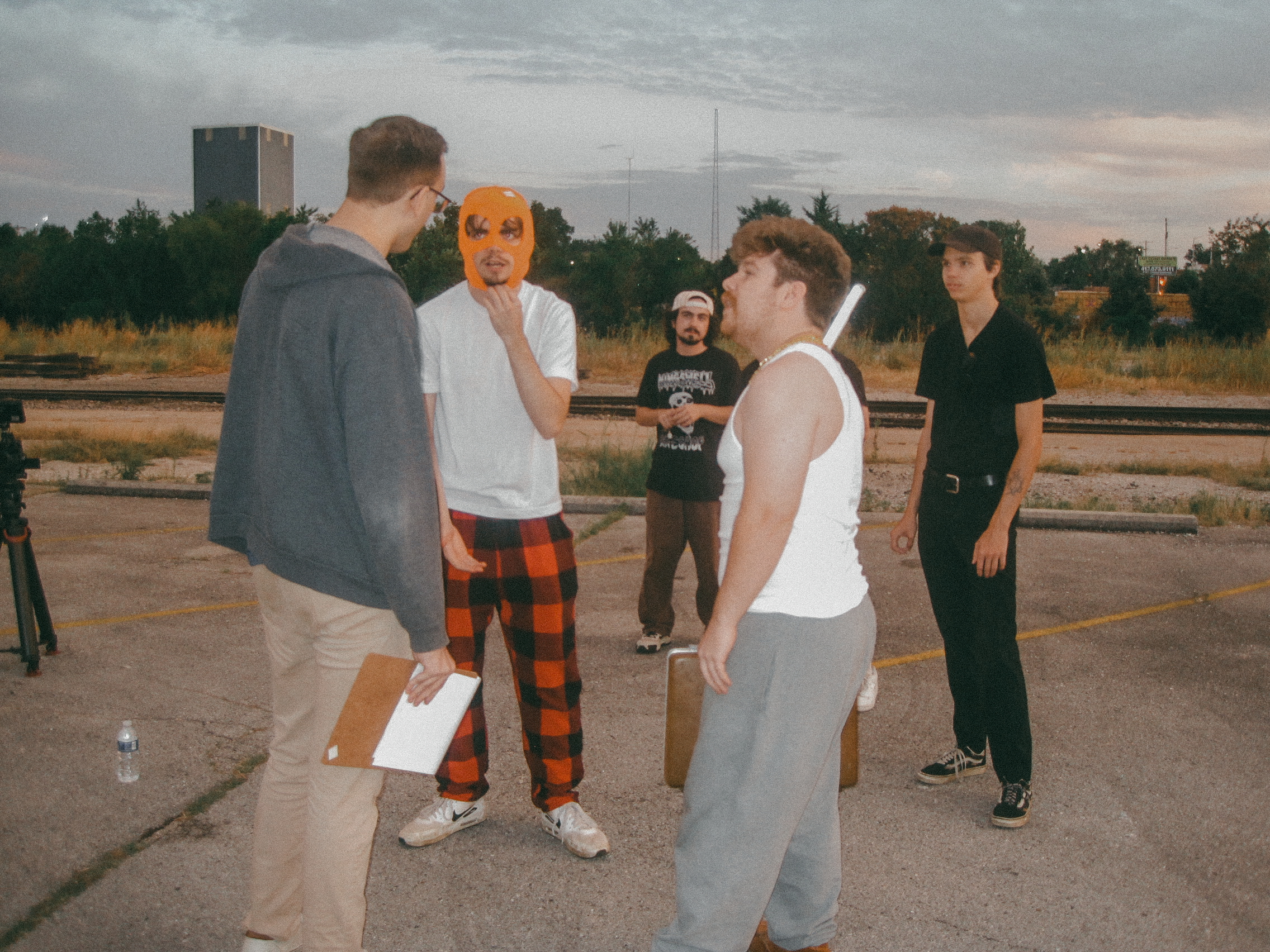 A group of five young people standing outdoors on a dirt or gravel surface, engaged in a conversation, with trees, a building, and railway tracks in the background during dusk or early evening.