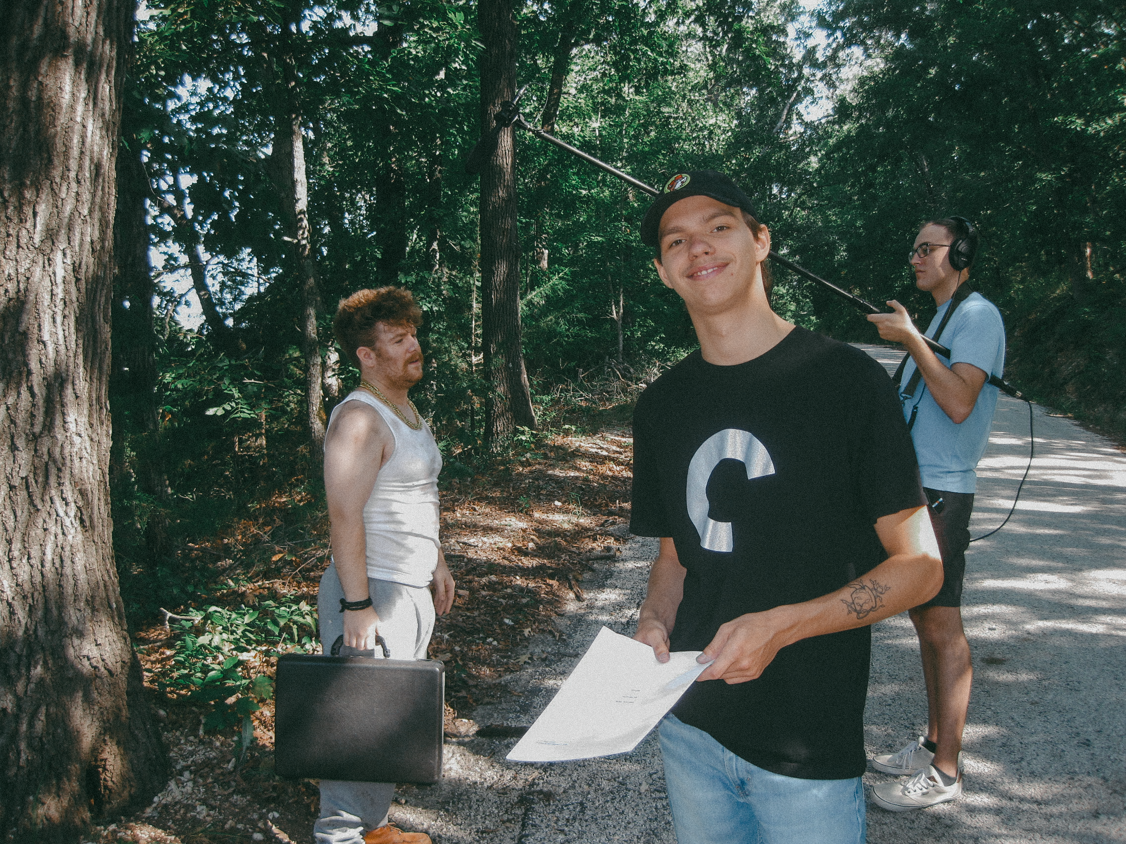 Three young men standing outdoors on a shaded wooded path, with one holding a sheet of paper and a third with headphones and a boom microphone assisting with audio recording.