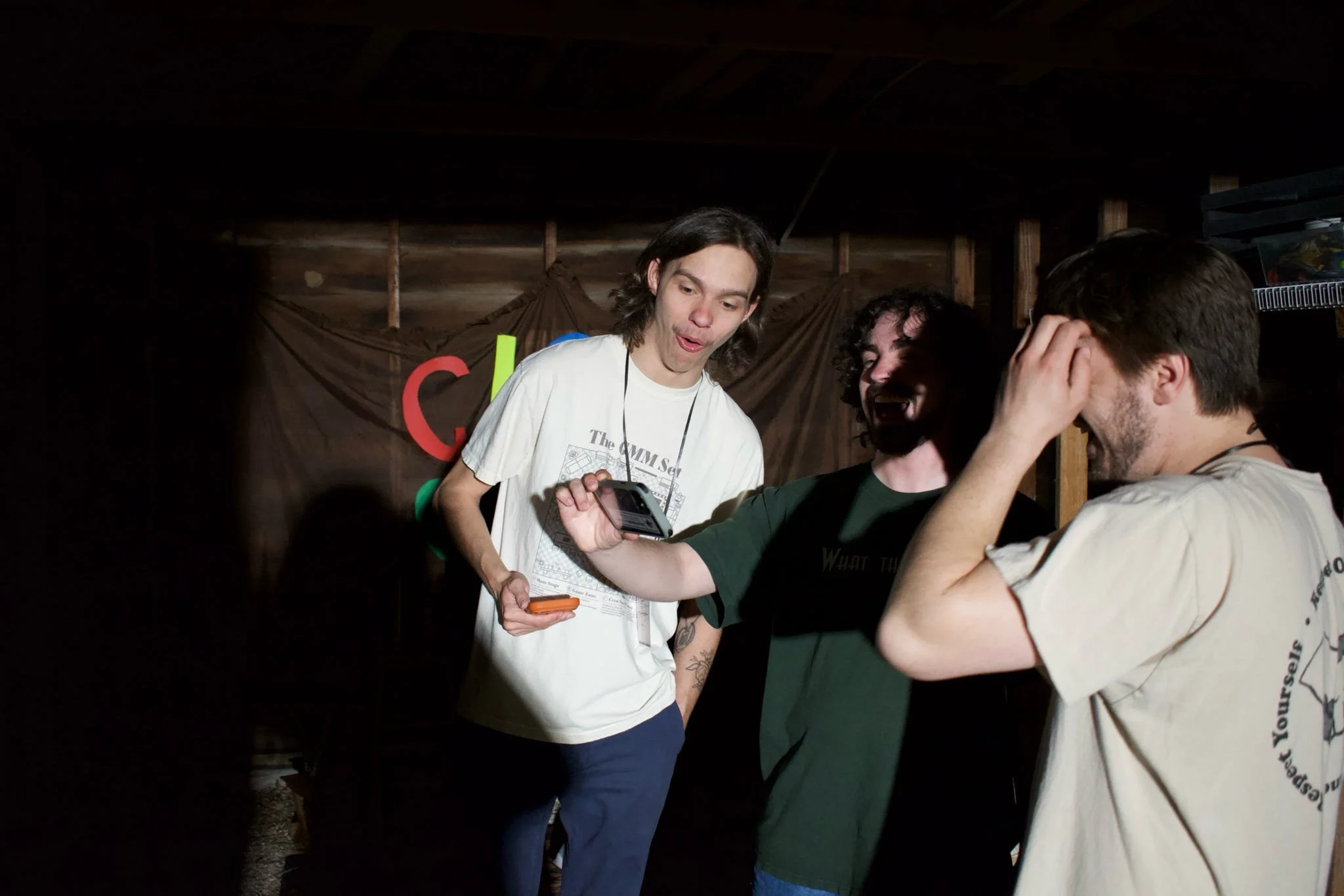 Three young men interacting in a dimly lit indoor space, with one holding a phone and the others reacting with surprise or amusement, and a dark background with a banner.