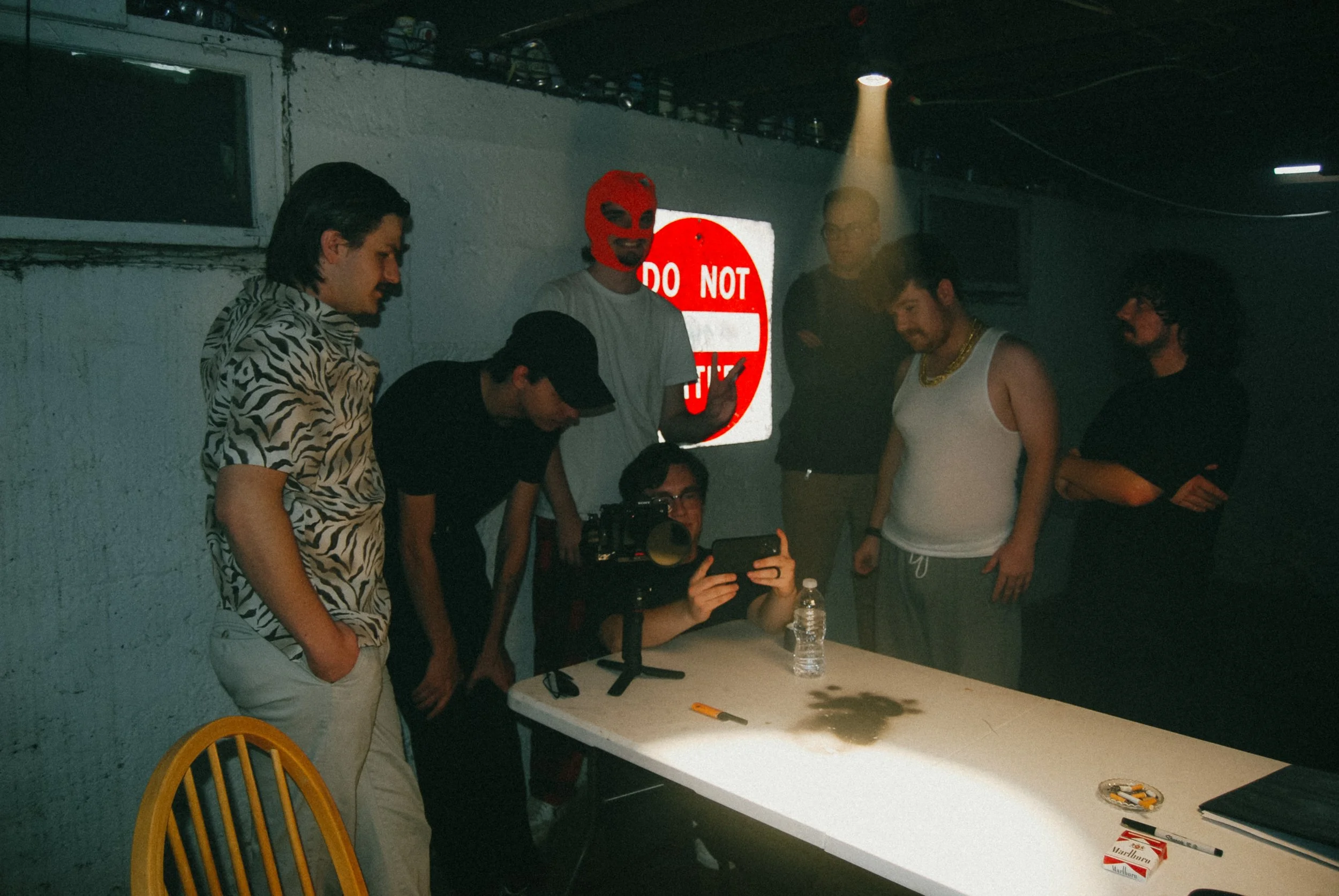 Group of young men gathered around a white table in a dimly lit room, some looking at a camera or phone, one wearing a red ski mask, and a