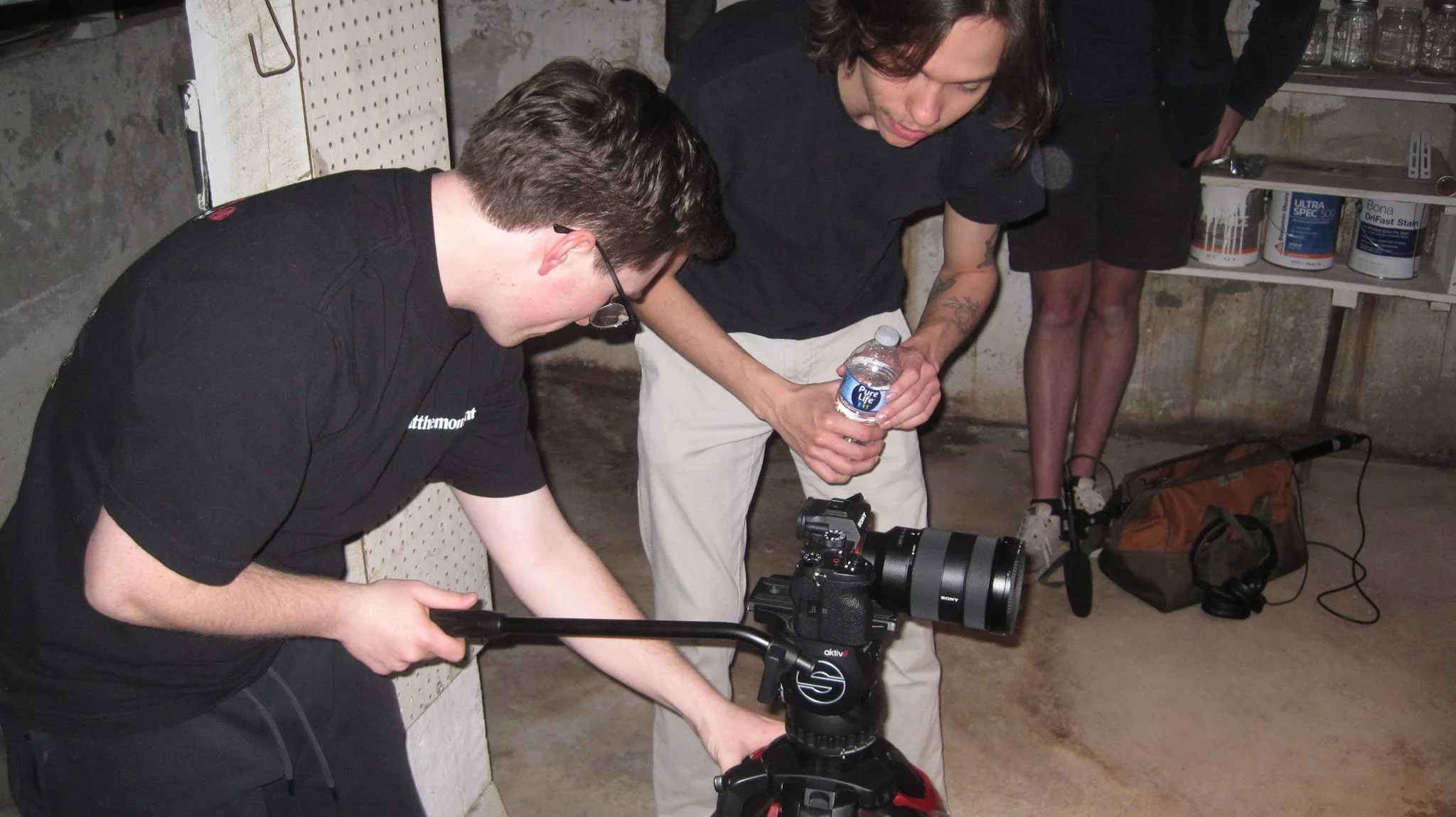Two men working with camera equipment in a basement or underground setting. One man is adjusting a camera, while the other holds a bottle of water. In the background, a woman in black shorts and a black shirt stands near shelves with paint cans.