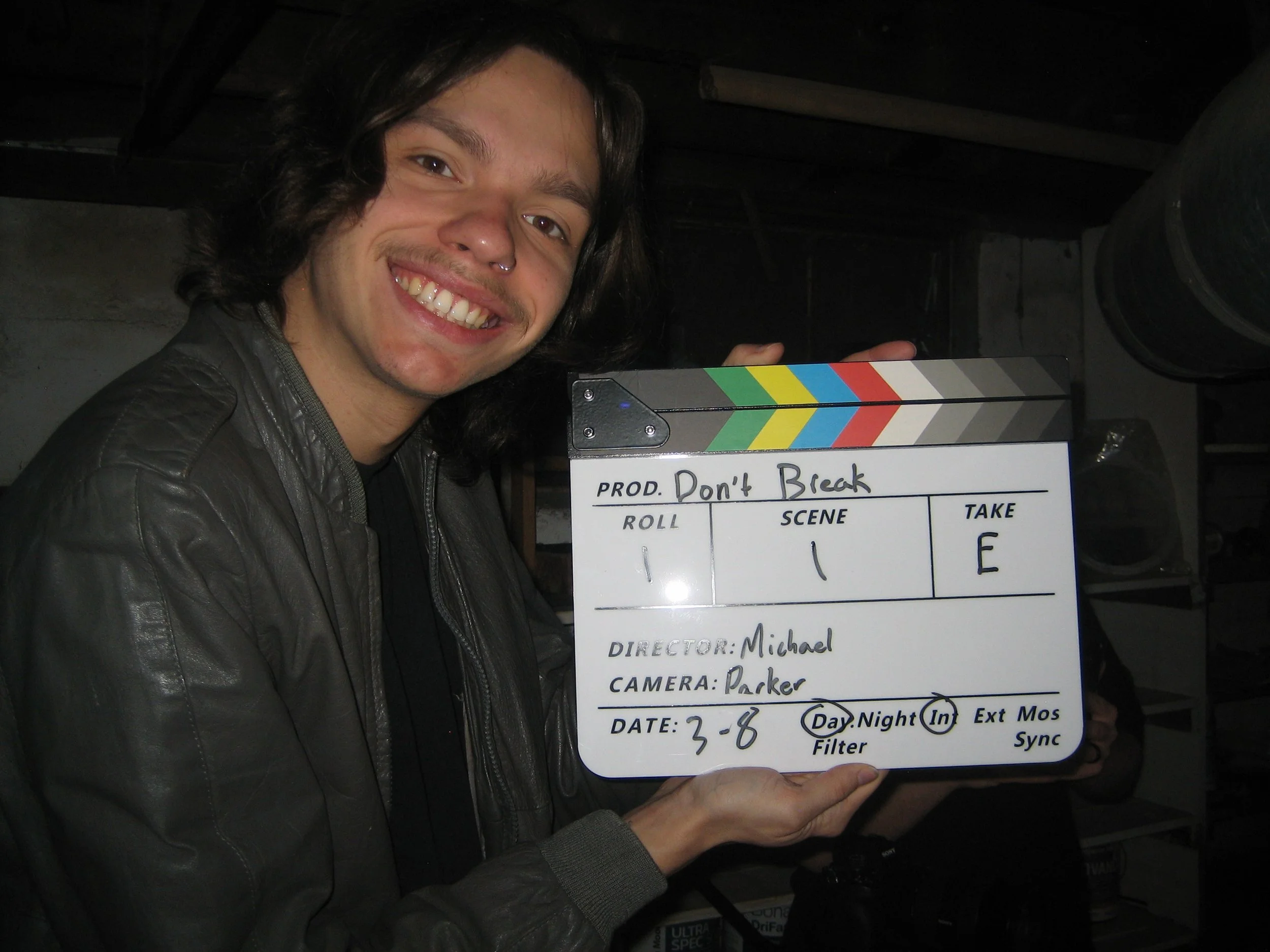 A person with long dark hair and a septum piercing smiling and holding a clapperboard with production details.