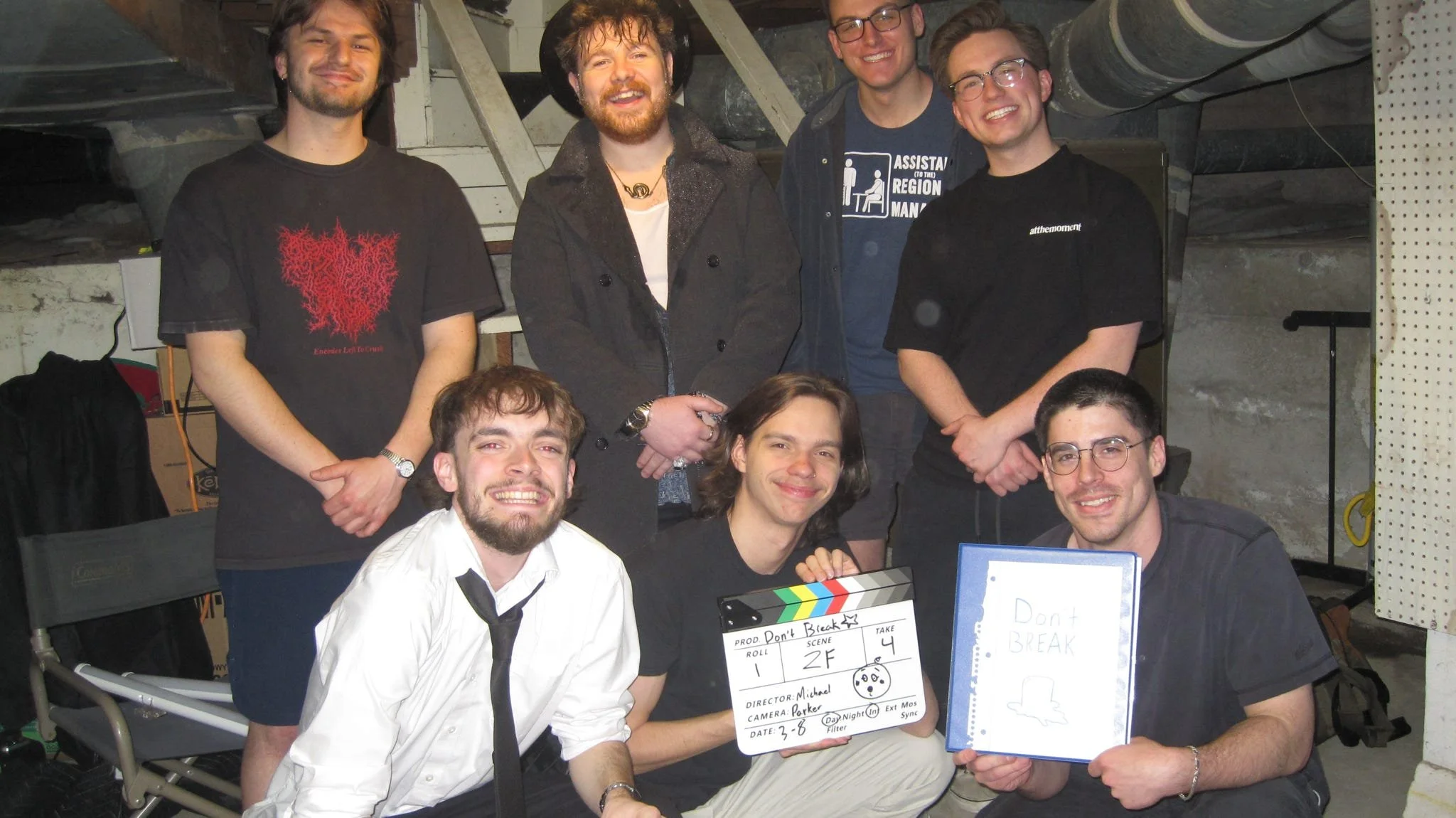 Group of six young men gathered together, smiling and posing for a photo in an indoor setting, possibly a basement or studio, with exposed concrete walls and ceiling pipes. They are casually dressed, with some wearing glasses and one wearing a hat. One person is holding a director's clapboard and another has a whiteboard with writing. There is filming equipment and a laptop visible in the scene.