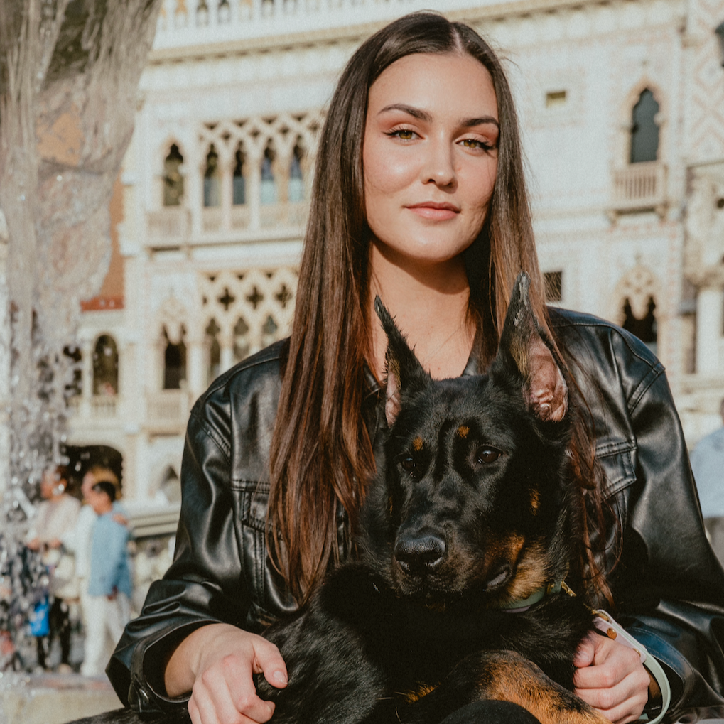 A young woman with long brown hair, wearing a black leather jacket, is holding a black and tan German Shepherd puppy outdoors in front of a historic, ornate building with arched windows and decorative stonework.