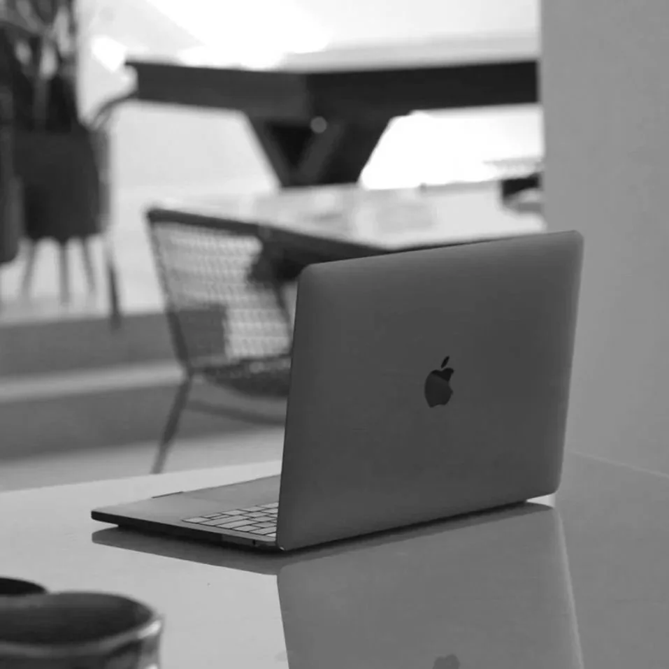A MacBook laptop on a desk with a chair and a dining table with chairs in the background.