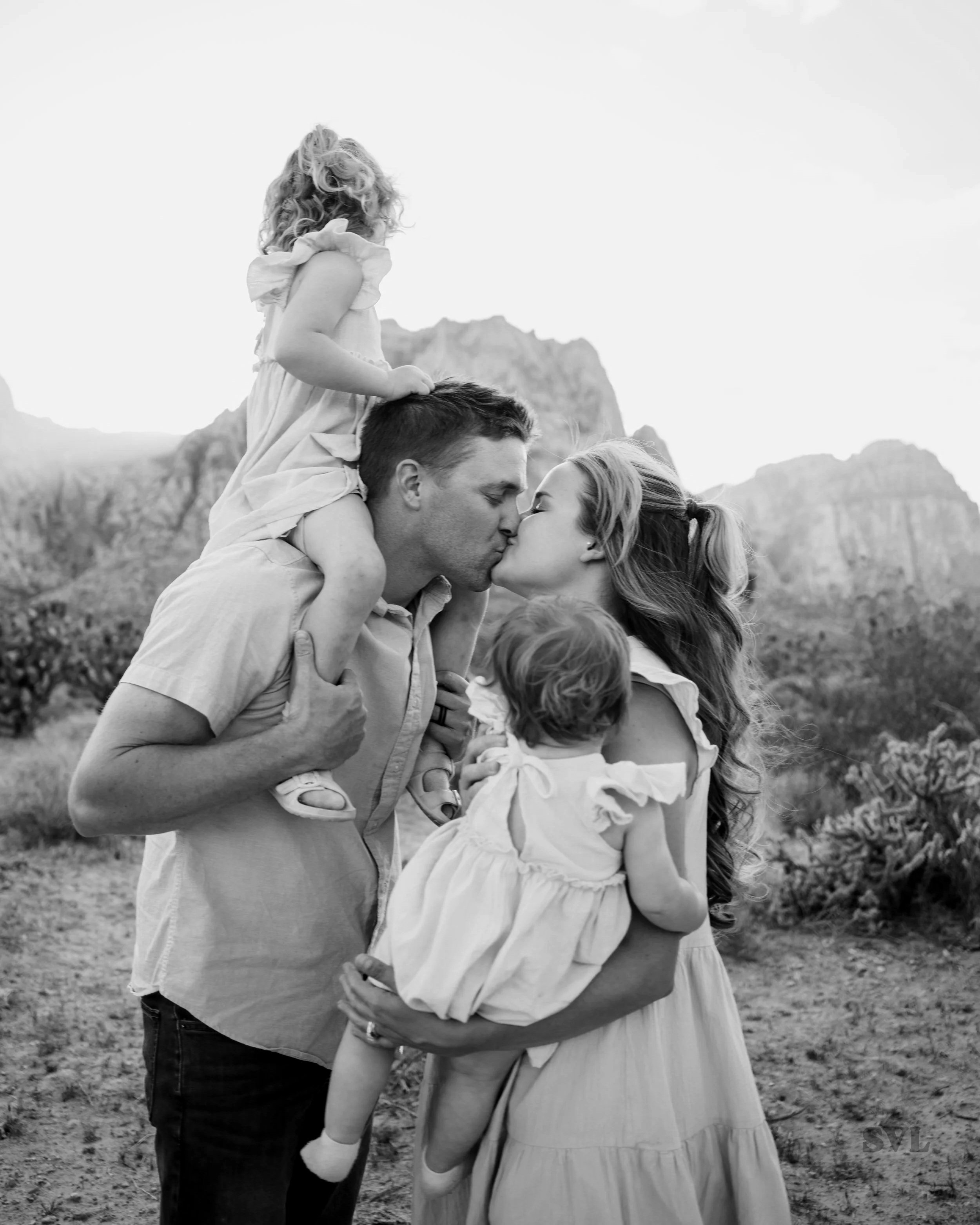 A black-and-white photo of a couple sharing a kiss in an outdoor setting with mountains in the background. A young girl sits on the man's shoulders, looking away, with her hair curly and wearing a dress with ruffled sleeves, while the woman has long hair and is wearing a light-colored dress.