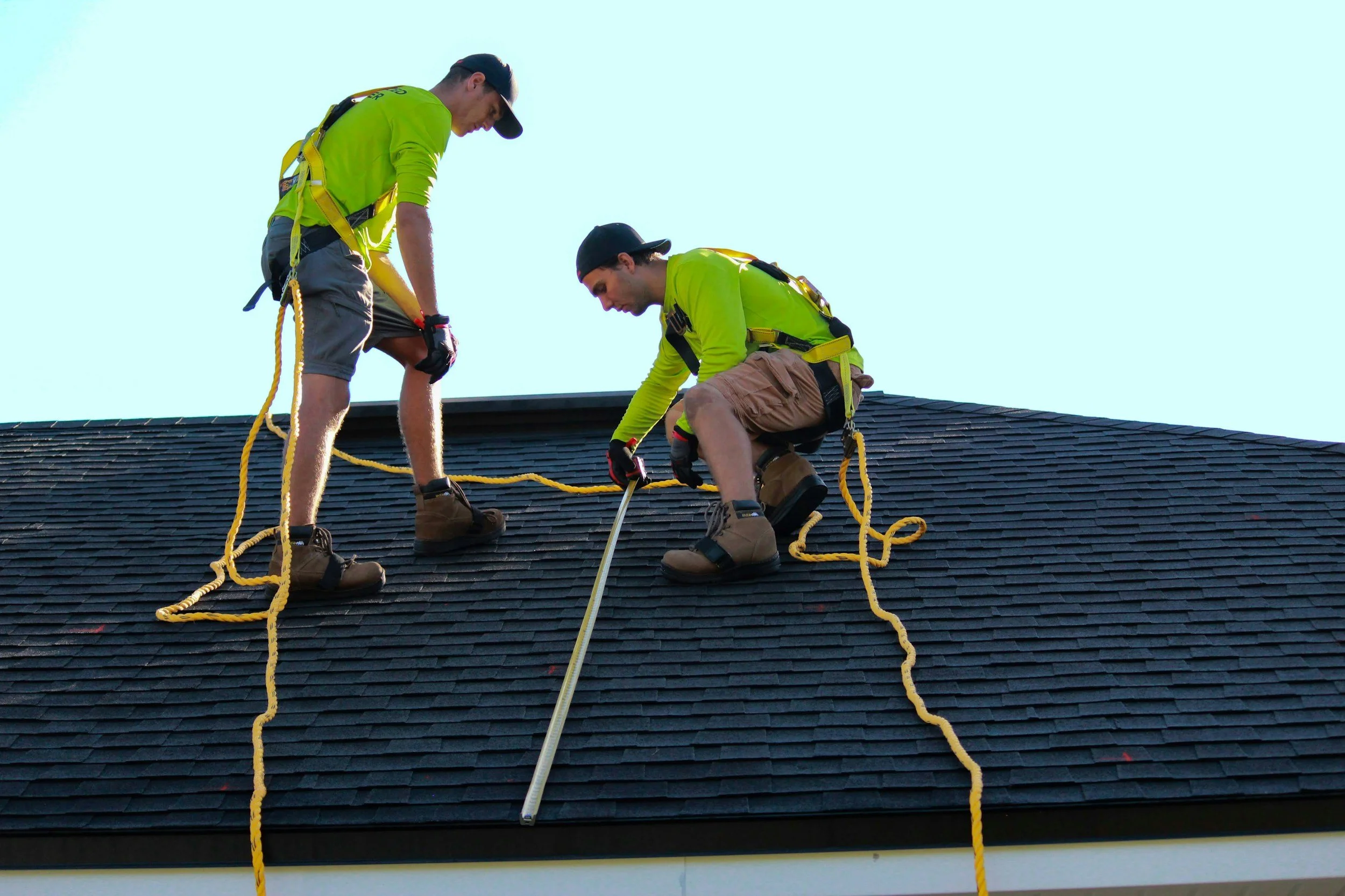 Two workers wearing safety harnesses and gloves, kneeling on a roof with black shingles, performing roof maintenance or repairs.