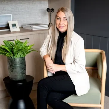 A woman with long blonde hair sitting on a light green chair, smiling, in a modern office or living space with a gray wall, a wooden cabinet, a potted plant, and some framed pictures in the background.