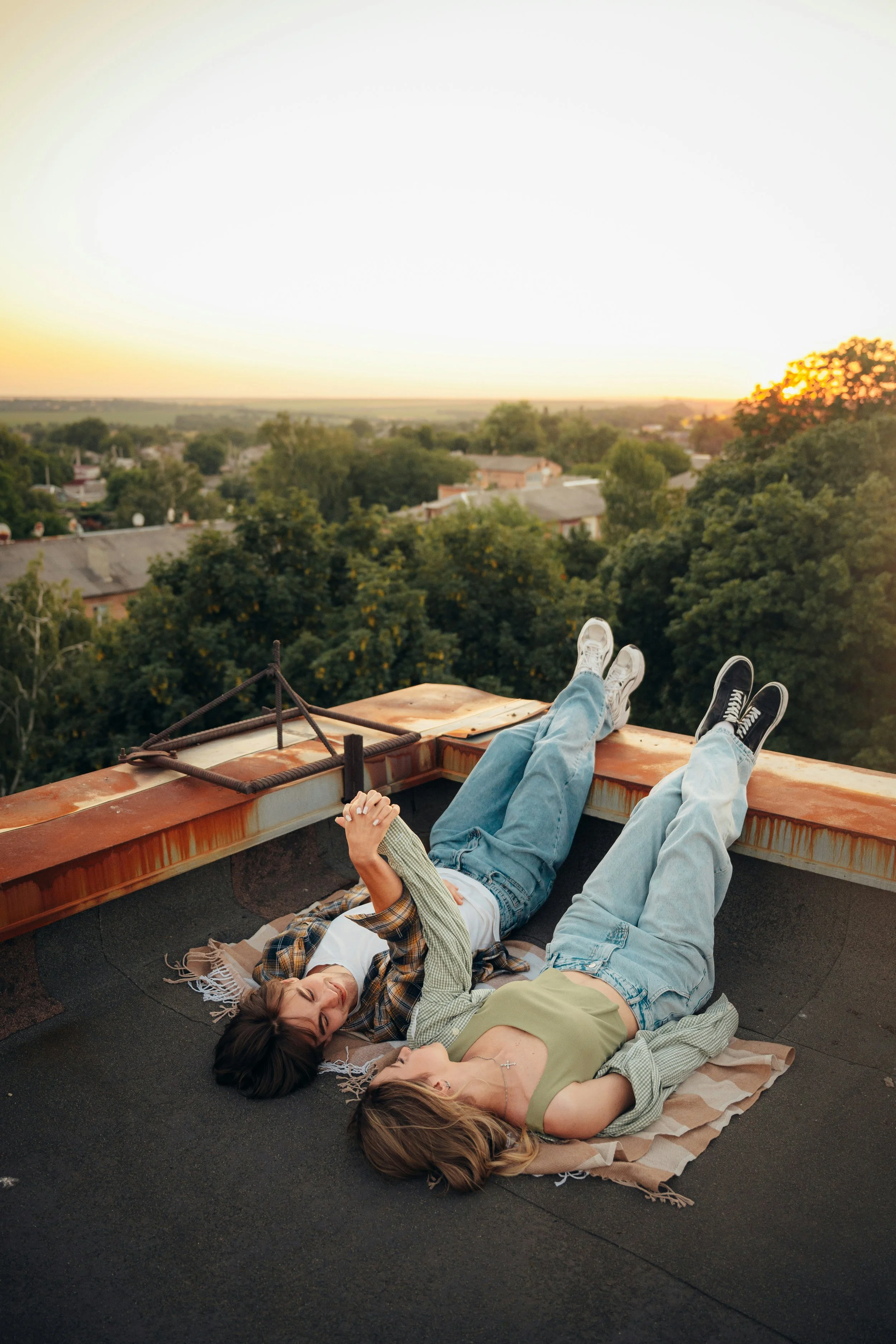 Two young women lying on a rooftop with city and trees in the distance at sunset, relaxing and holding hands.