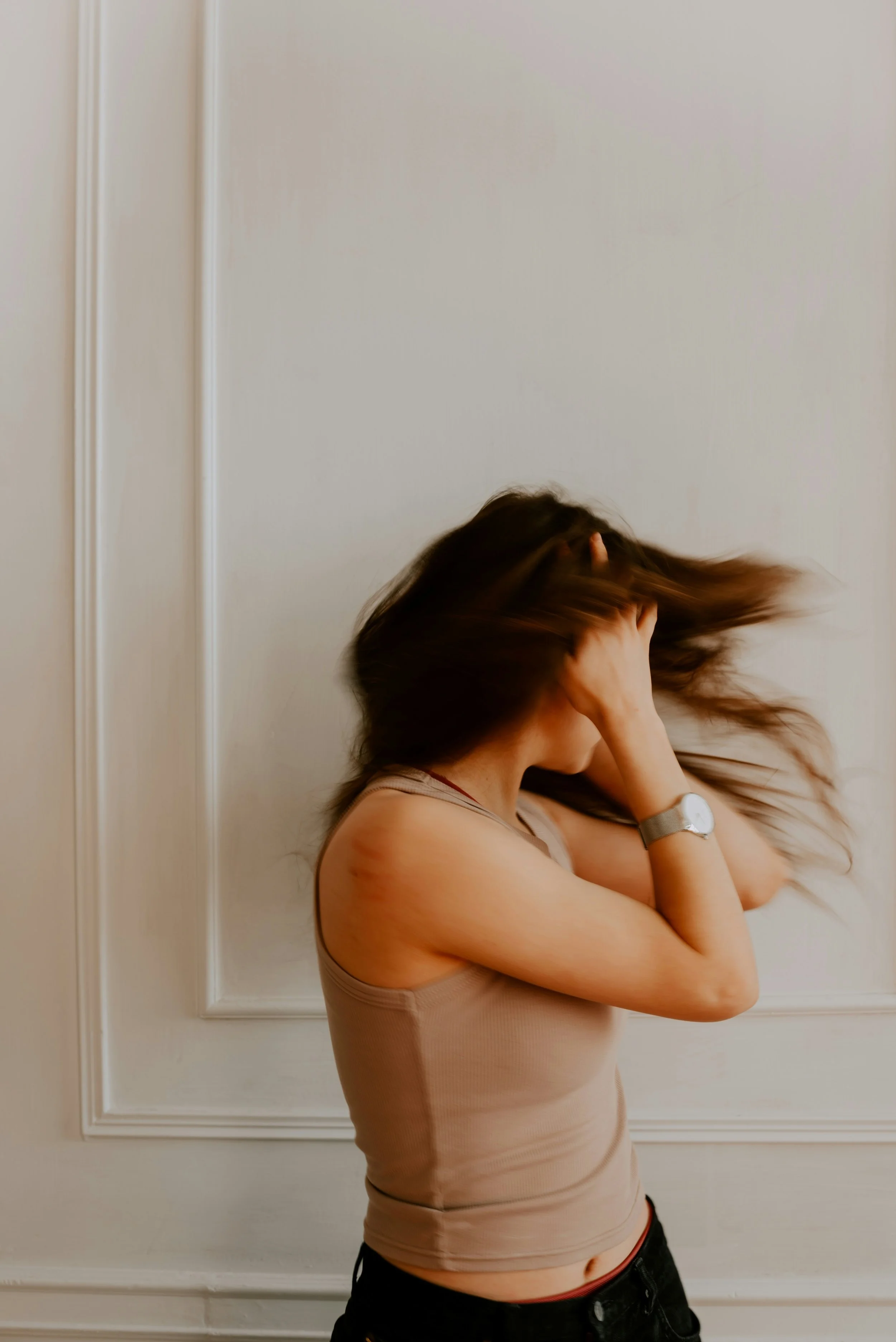 A woman with brown hair wearing a beige tank top and black pants, standing against a white wall with decorative molding, flipping her hair with her head tilted downward.