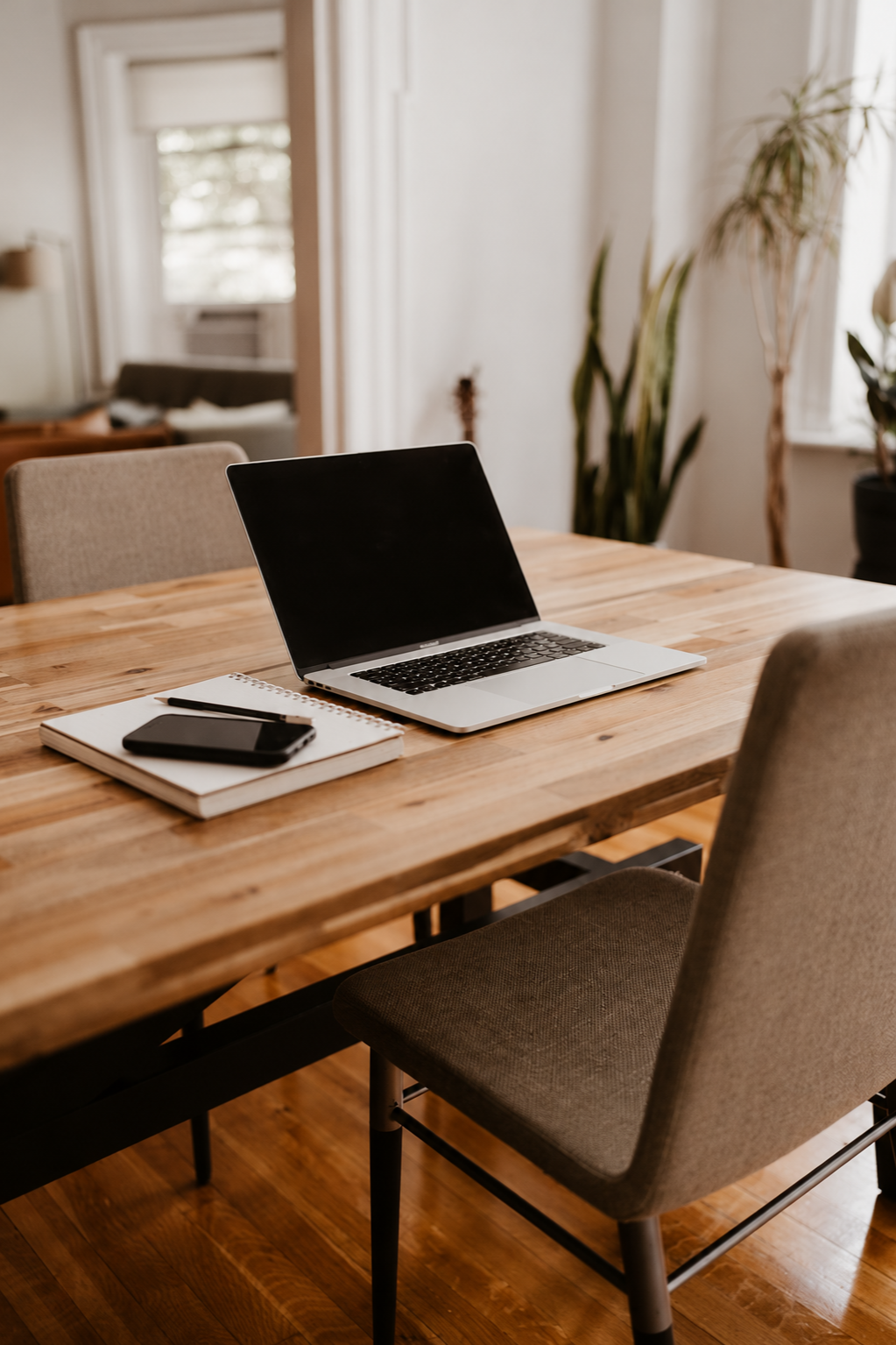 Work space with a wooden desk, a laptop, a notebook, a pen, and a smartphone, surrounded by chairs and green plants in a bright room.